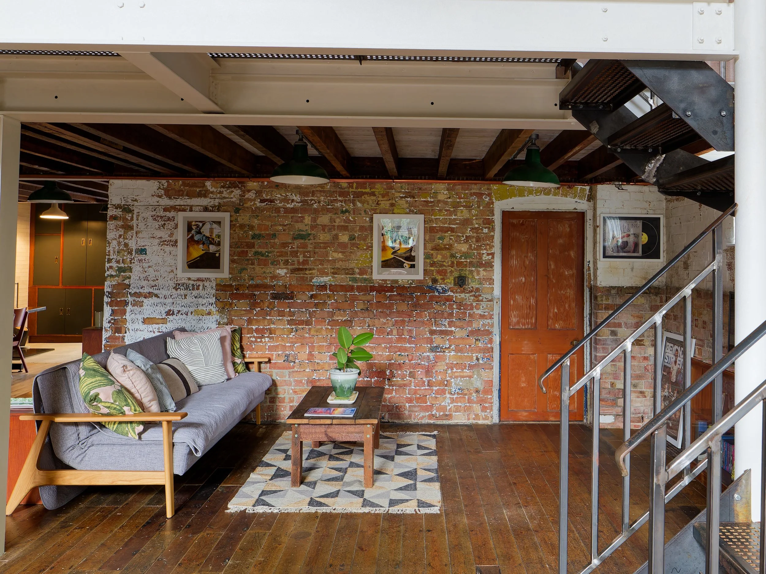 Living room with exposed brick wall, wooden floor, grey sofa with patterned pillows, small wooden coffee table with a potted plant, framed art on brick wall, wooden door, staircase with metal railing, and ceiling with green pendant lights.