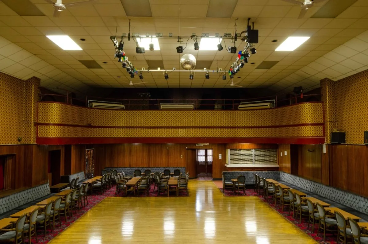 An empty banquet hall or event venue with wooden floors, decorated walls with gold and red patterns, a central dance floor, tables and chairs arranged along the sides, a balcony with seating, and stage lighting equipment hanging from the ceiling.