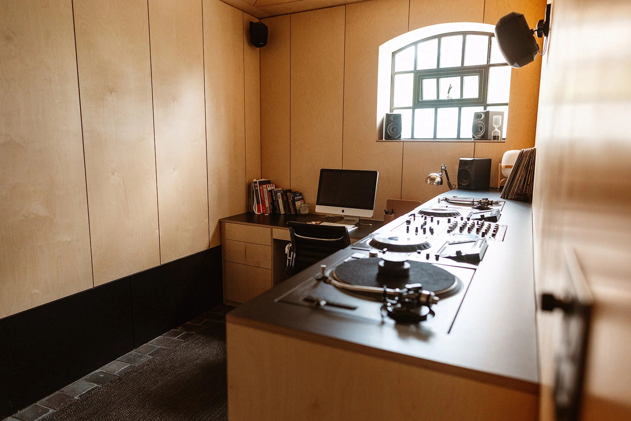An indoor DJ studio with a wooden wall, computer, books, and DJ equipment including turntables, mixer, and speakers near a window.