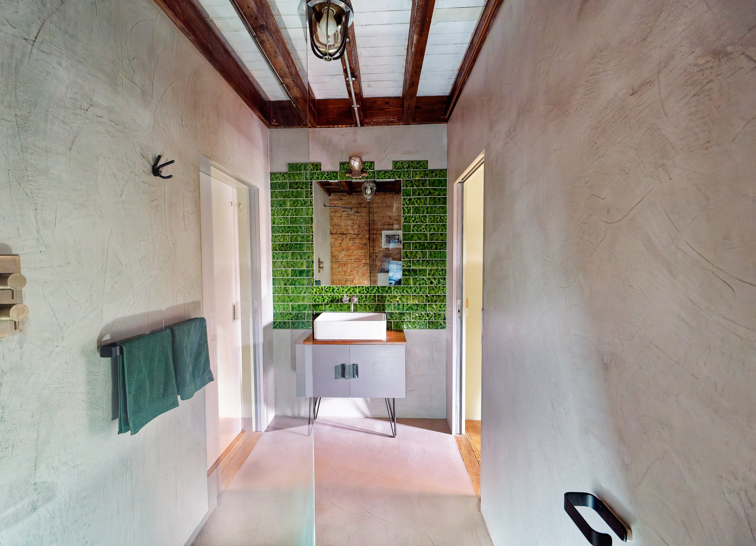A bathroom with a textured beige wall on the right and a green tiled wall behind a white rectangular sink with a wooden countertop. Above the sink is a large square mirror, and on the wall to the left is a towel rack with green towels. The ceiling ha