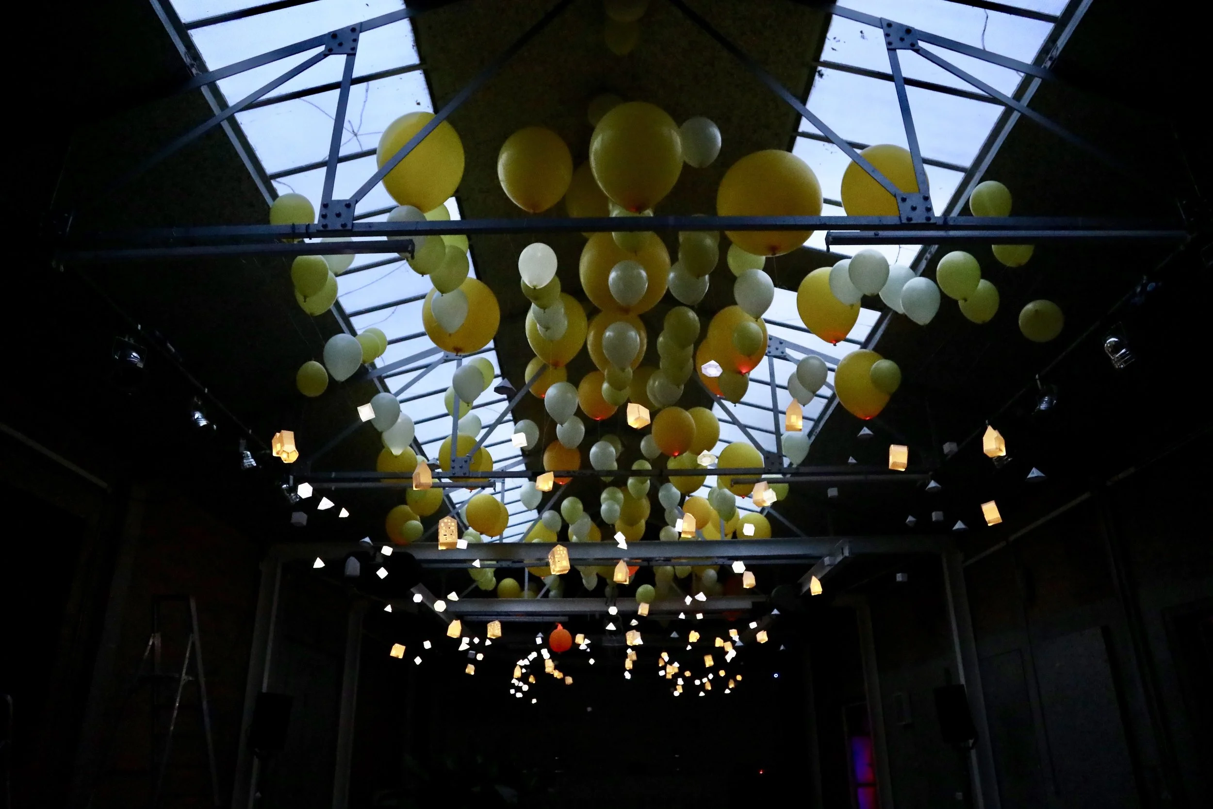 Ceiling installation with yellow and white balloons hanging from a framework, with small hanging lanterns and light fixtures illuminating a dark room, with skylight windows above.