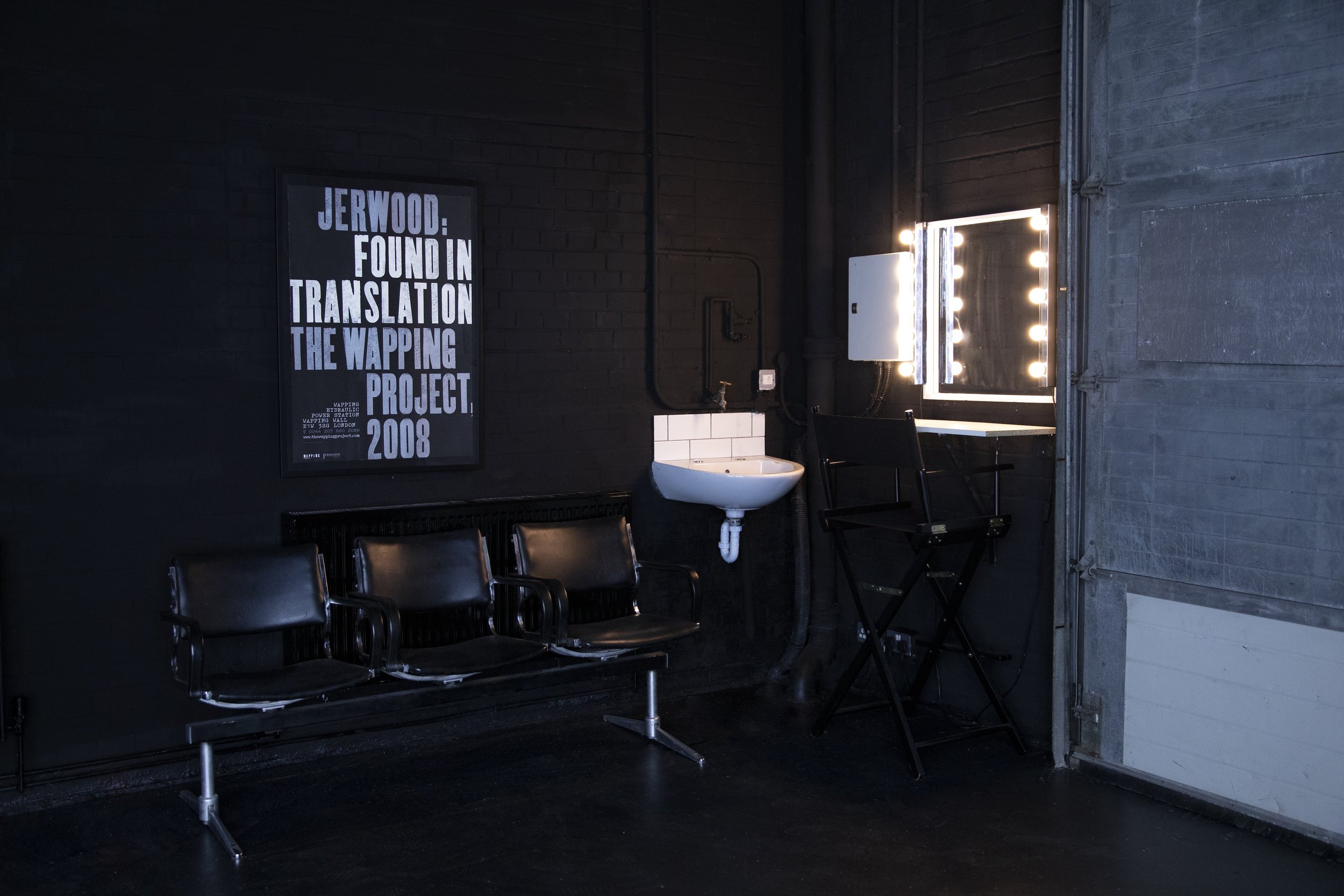 Interior of a room with black walls, containing black seated chairs, a small white sink, a black chair, and a makeup mirror with light bulbs, next to a window with closed blinds and a poster on the wall.