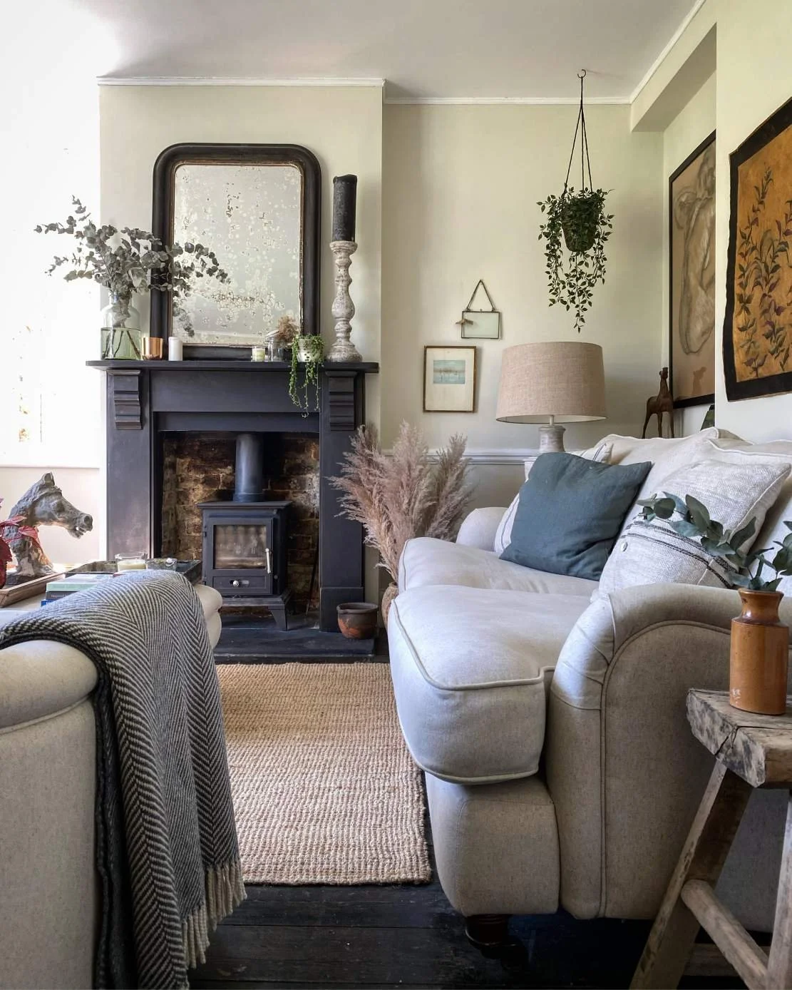 Cozy living room with a cream sofa, decorative pillows, and a side table with a vase. A black fireplace mantel with a mirror, candles, and plants. Wall art, hanging plants, and a beige area rug on dark wooden floors.