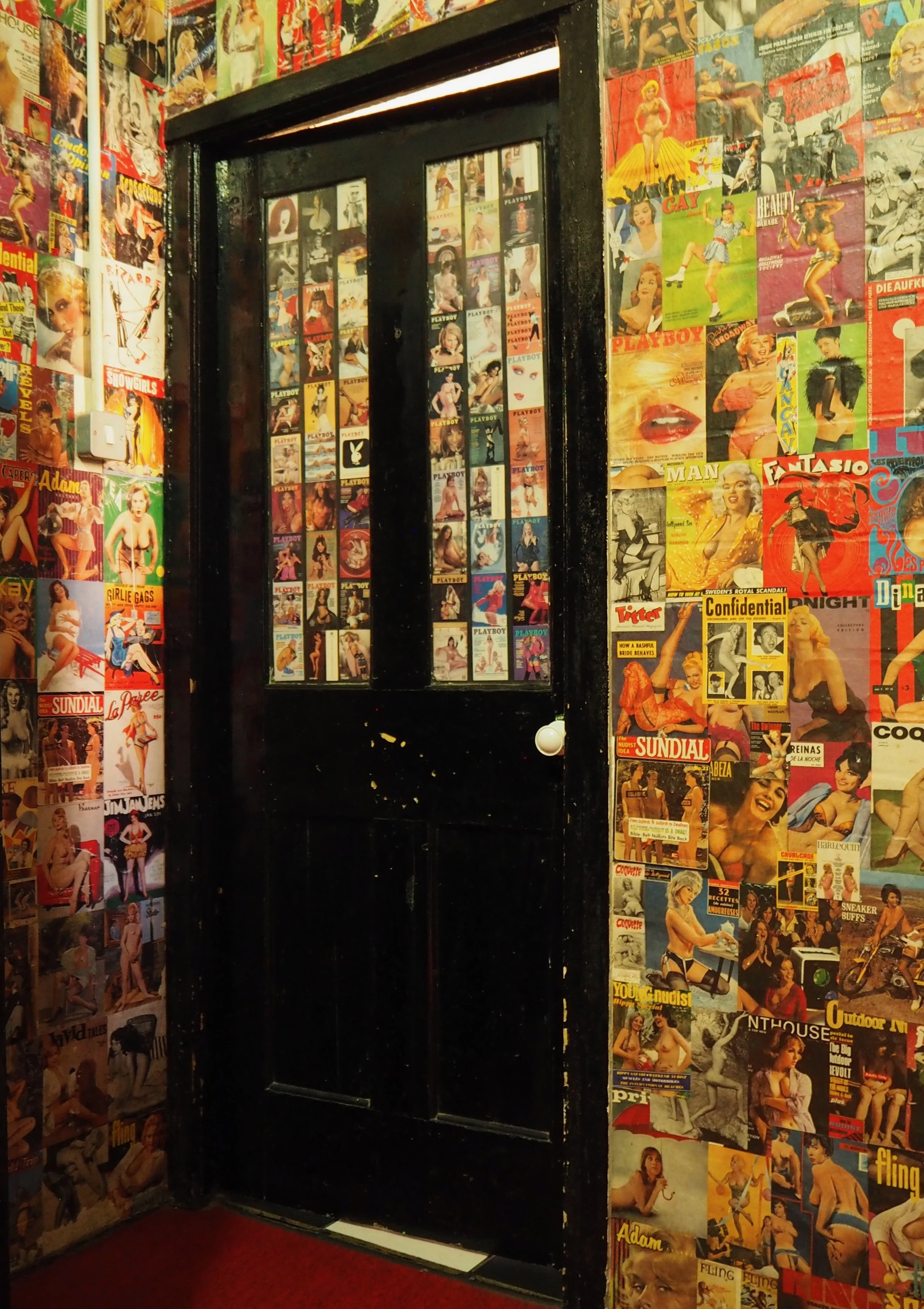 A black door decorated with numerous vintage Playboy magazine covers, surrounded by walls covered with similar magazine covers.