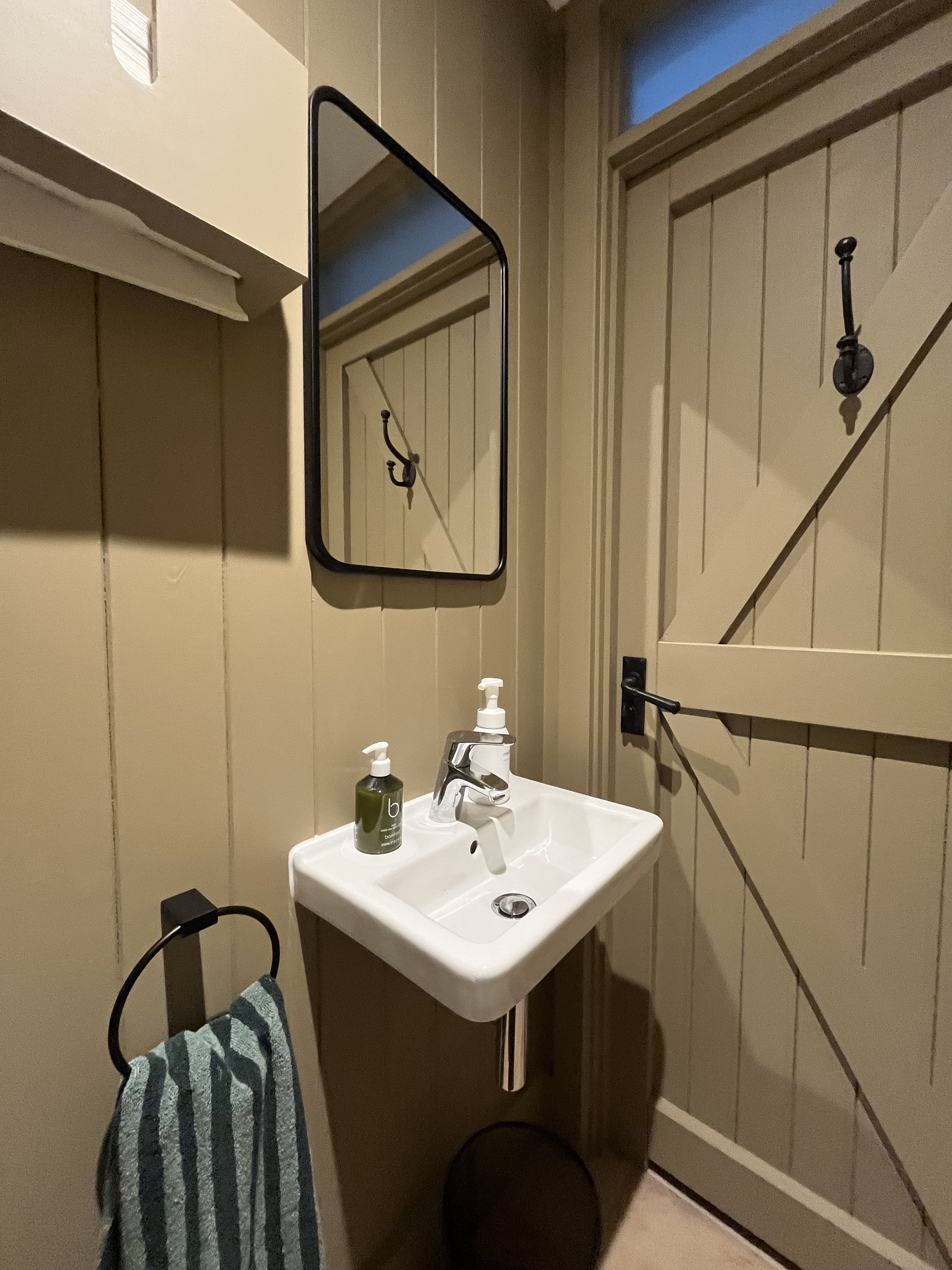 A small bathroom with beige wooden paneled walls, a white wall-mounted sink with a chrome faucet, a soap dispenser, a towel hanging on a black ring holder, a black-framed mirror above the sink, a black hook on the wall, and a beige wooden door with d