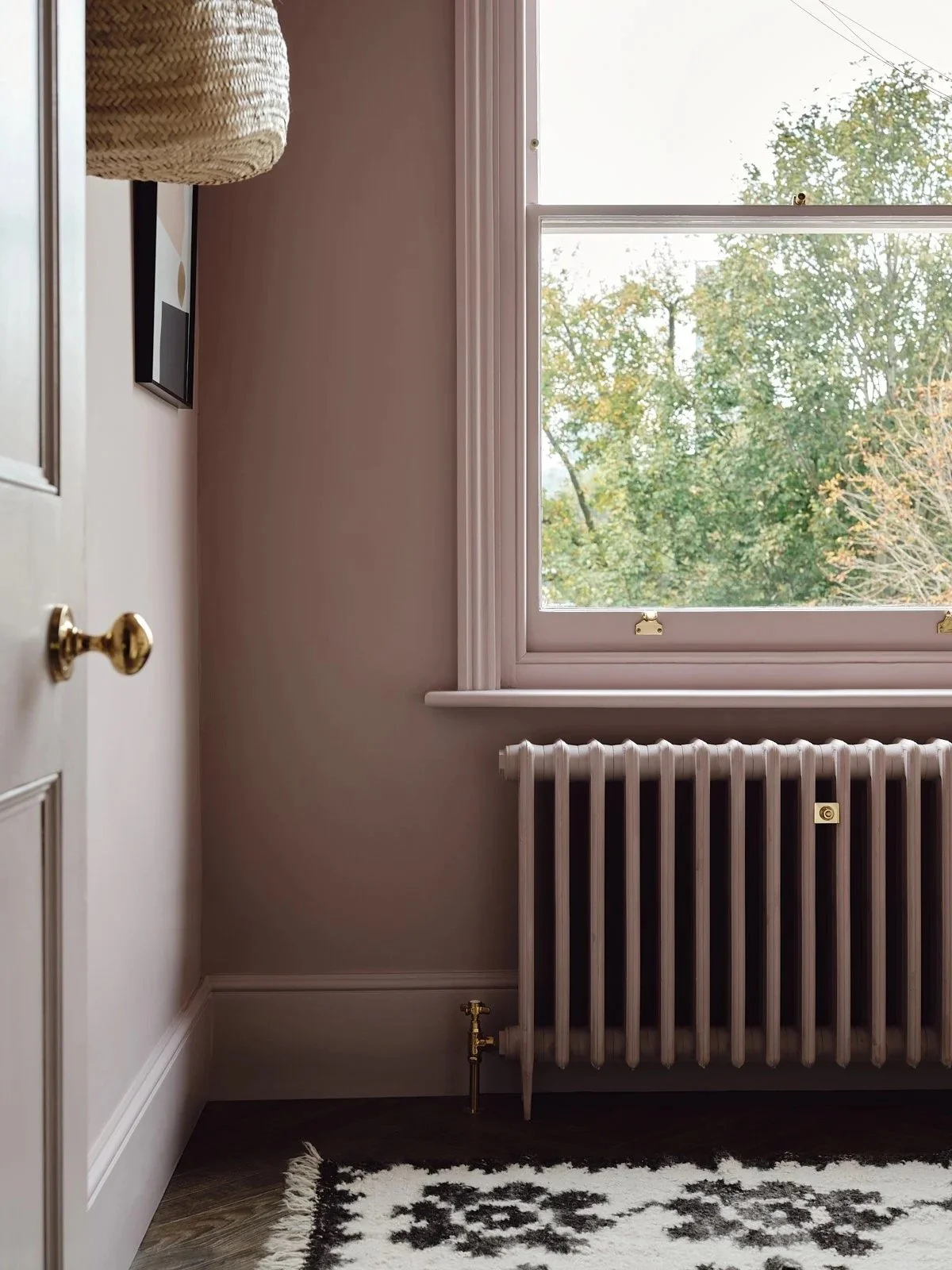 A corner of a room showing a window with trees outside, a white radiator below the window, a portion of a door on the left, and a black and white rug on a wooden floor