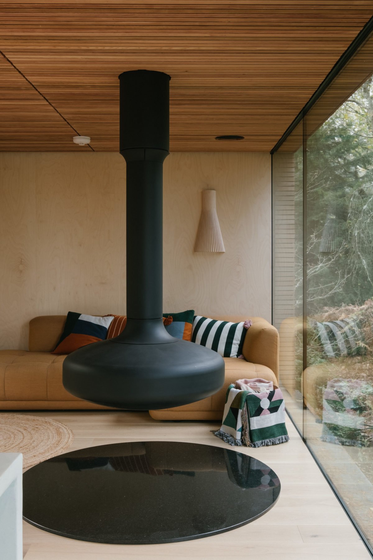 Interior of a modern living room with a beige sofa adorned with colorful pillows, a black suspended fireplace, a round black coffee table, and a large glass window showing a view of greenery outside, with wooden ceiling panels.