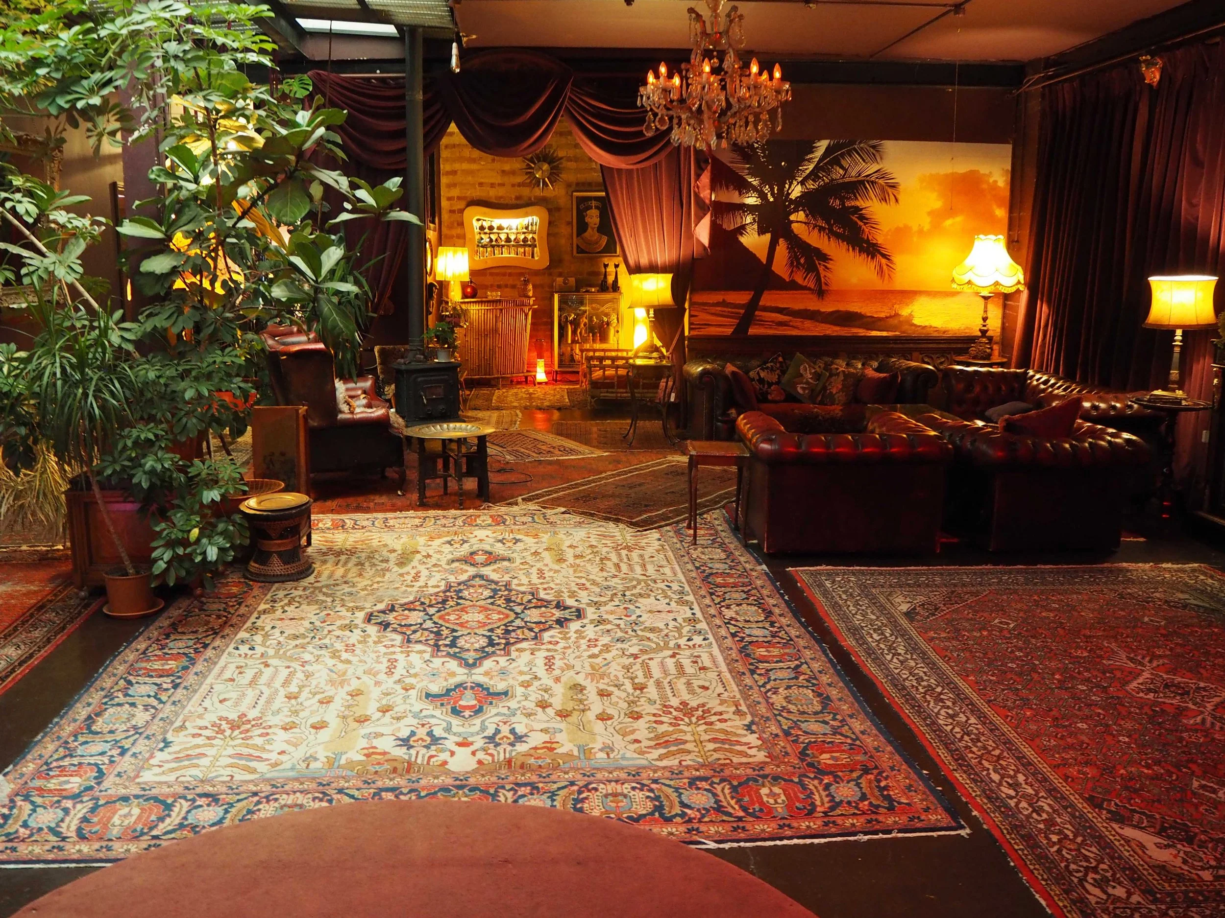 A cozy vintage living room with Persian rugs, leather sofas, lush plants, warm lighting, a chandelier, and a sunset beach mural on the wall.
