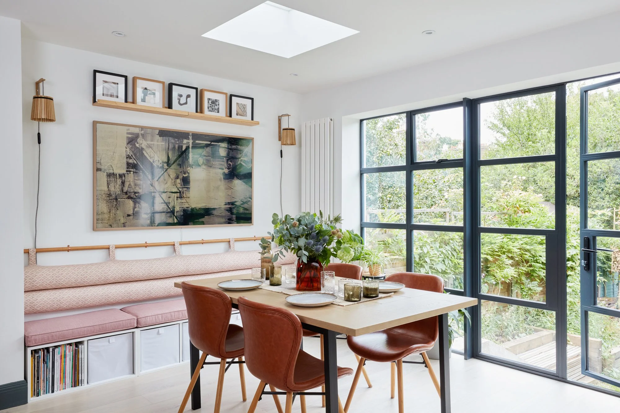 Modern dining room with large windows, a light wood table, four terracotta chairs, a built-in bench with storage, a large abstract painting, smaller framed artworks, houseplants, and a skylight.