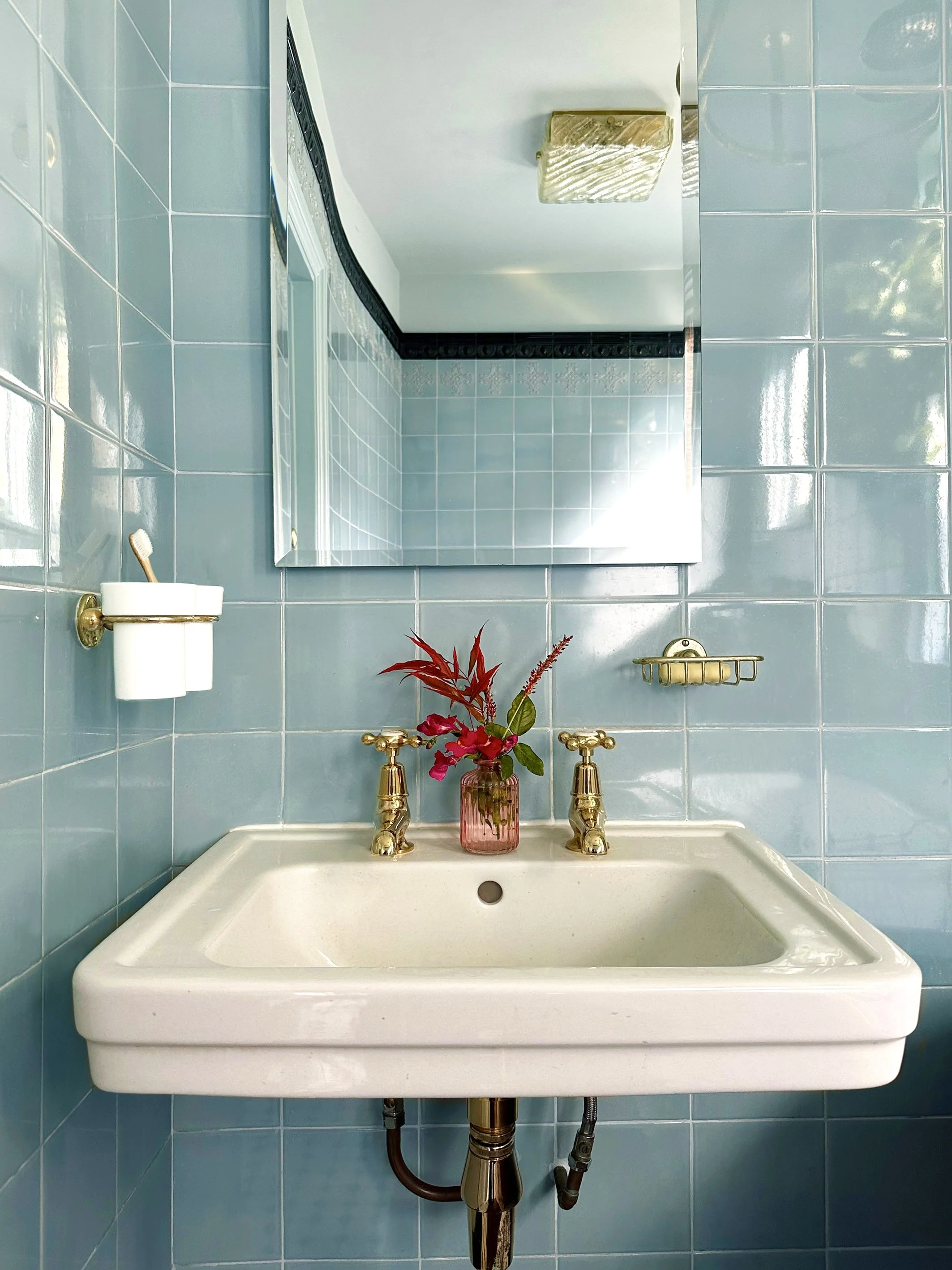 A vintage bathroom sink with a gold faucet, a pink vase with red and pink flowers, and a mirror on a blue tiled wall.