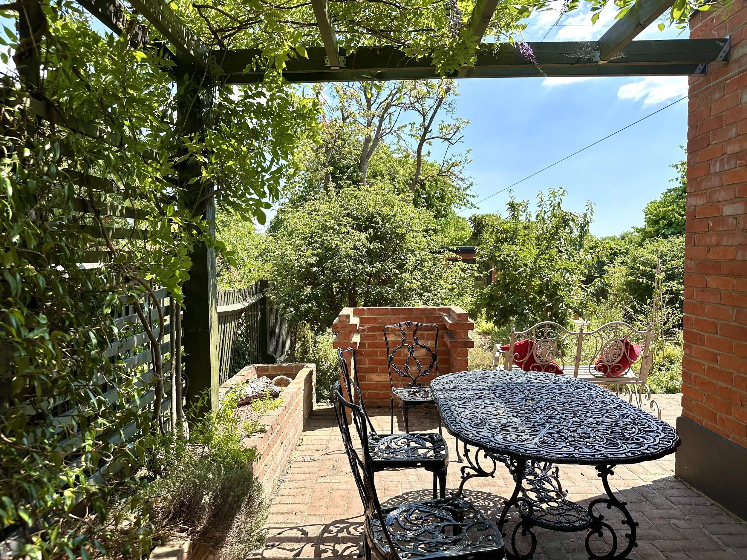 A cozy outdoor patio with a wrought iron table and chairs, surrounded by lush green plants and trees under a sunny sky.