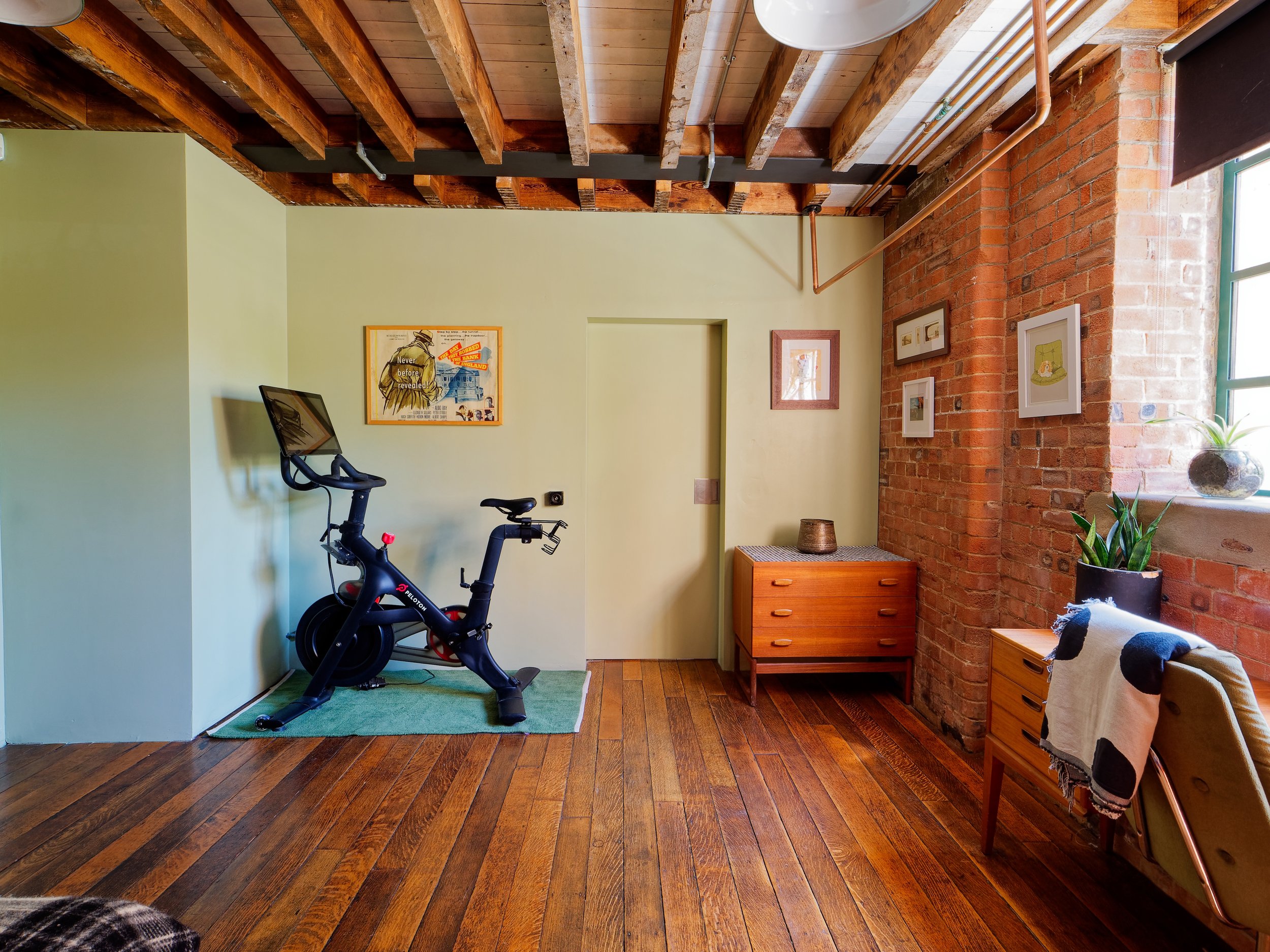 A room with wooden floors, exposed brick wall, and yellowish walls. There is a stationary exercise bike with a monitor and a small green rug underneath, placed against one wall. Decor includes framed pictures and artwork on the walls. A window with a