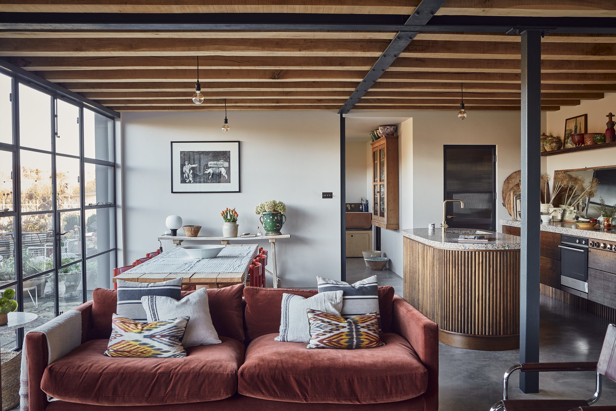Open-concept living and dining space with a kitchen visible in the background, large windows on the left, wooden ceiling beams, a red sofa with patterned pillows, and decorative items including plants and artwork.