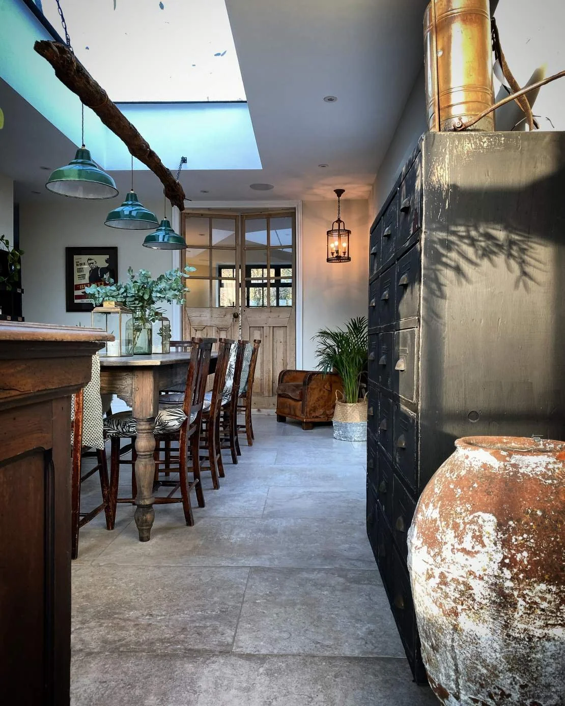 A cozy dining area with a rustic wooden table, chairs with patterned cushions, a glass vase with eucalyptus leaves, a hanging lantern, and a black cabinet. There's a large terracotta pot on the right and a potted plant near the back. The cabin-style 