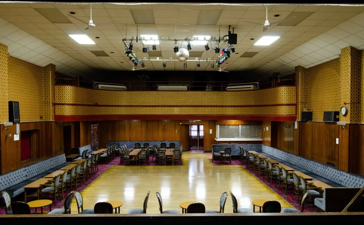 Empty banquet hall with a large open dance floor, surrounding tables, chairs, and an upper balcony with additional seating.