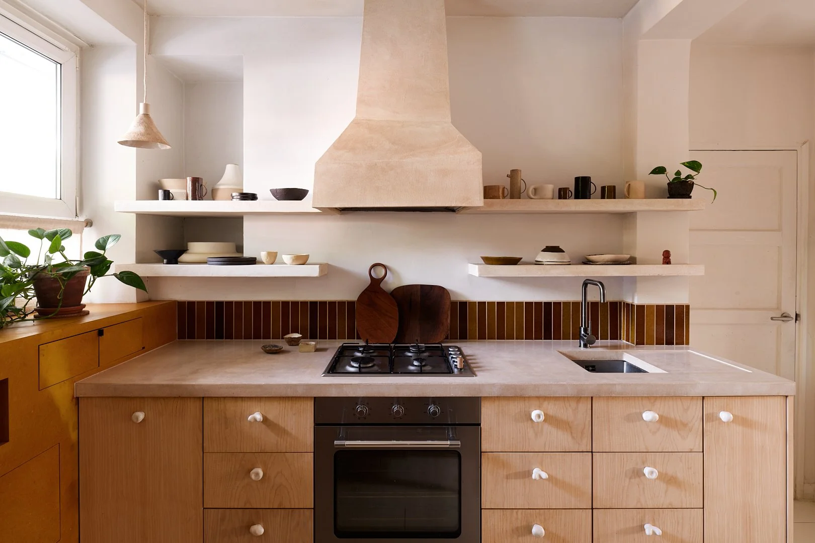 Modern kitchen with light wood cabinets, black oven, gas cooktop, small sink, white shelves with dishes and cups, potted plant near window, pendant light, beige range hood, decorative cutting boards, and small bowls.