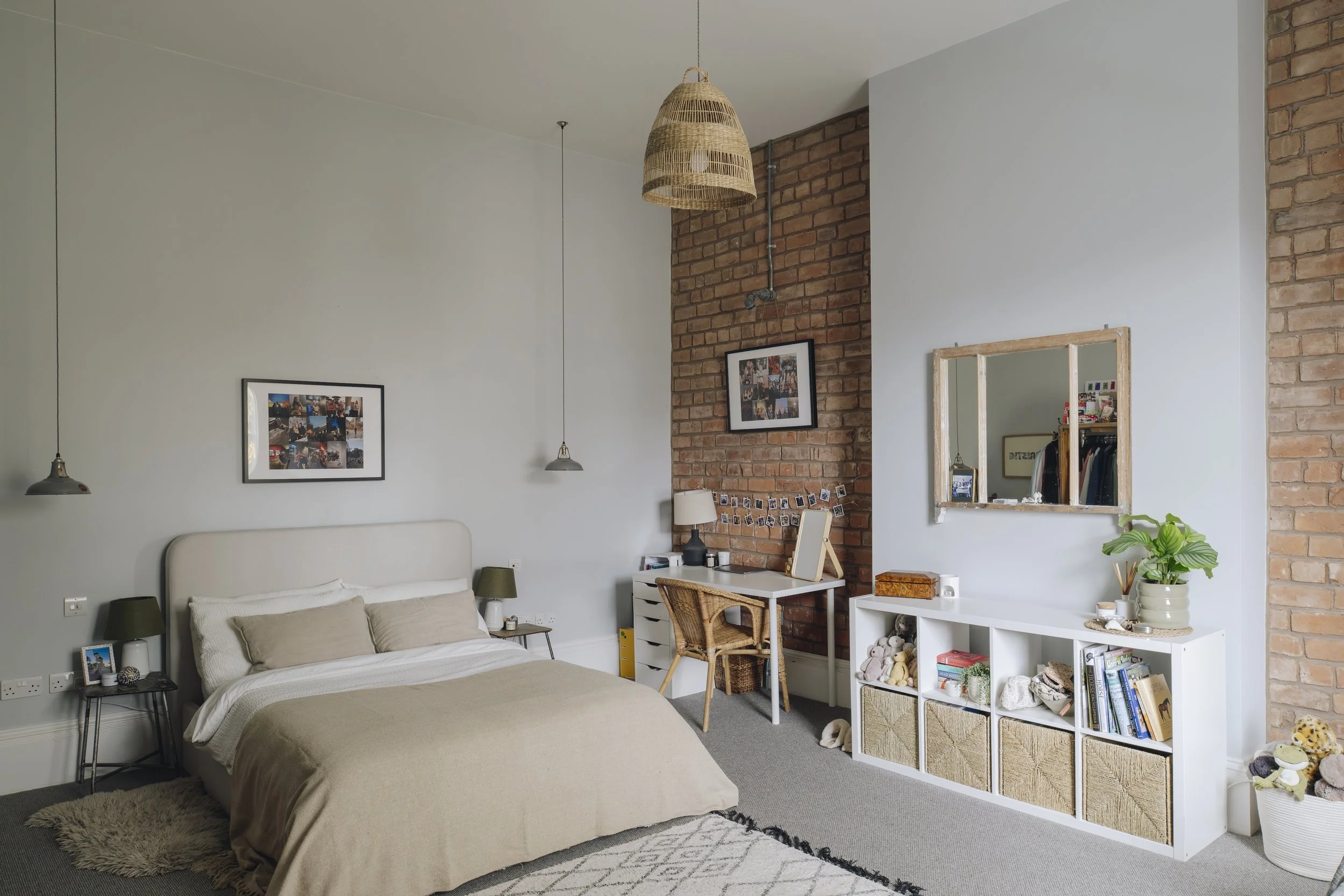 A bedroom with a bed having white and beige linens, two side tables with lamps, a brick accent wall, a desk with a wicker chair, a mirror, and shelves with books and toys.