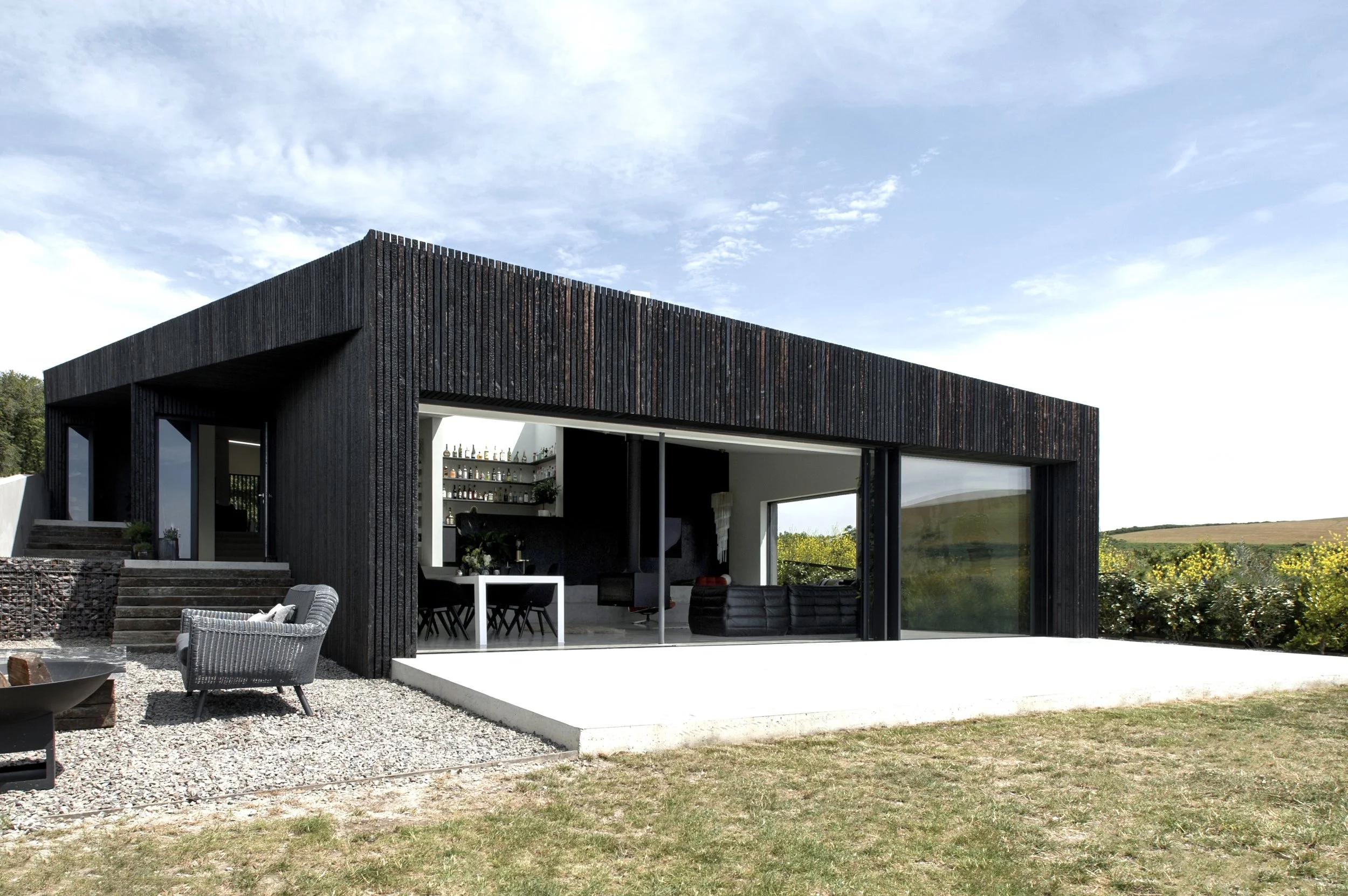 Modern black house with large glass sliding doors, outdoor seating area, and a view of distant fields under a partly cloudy sky.
