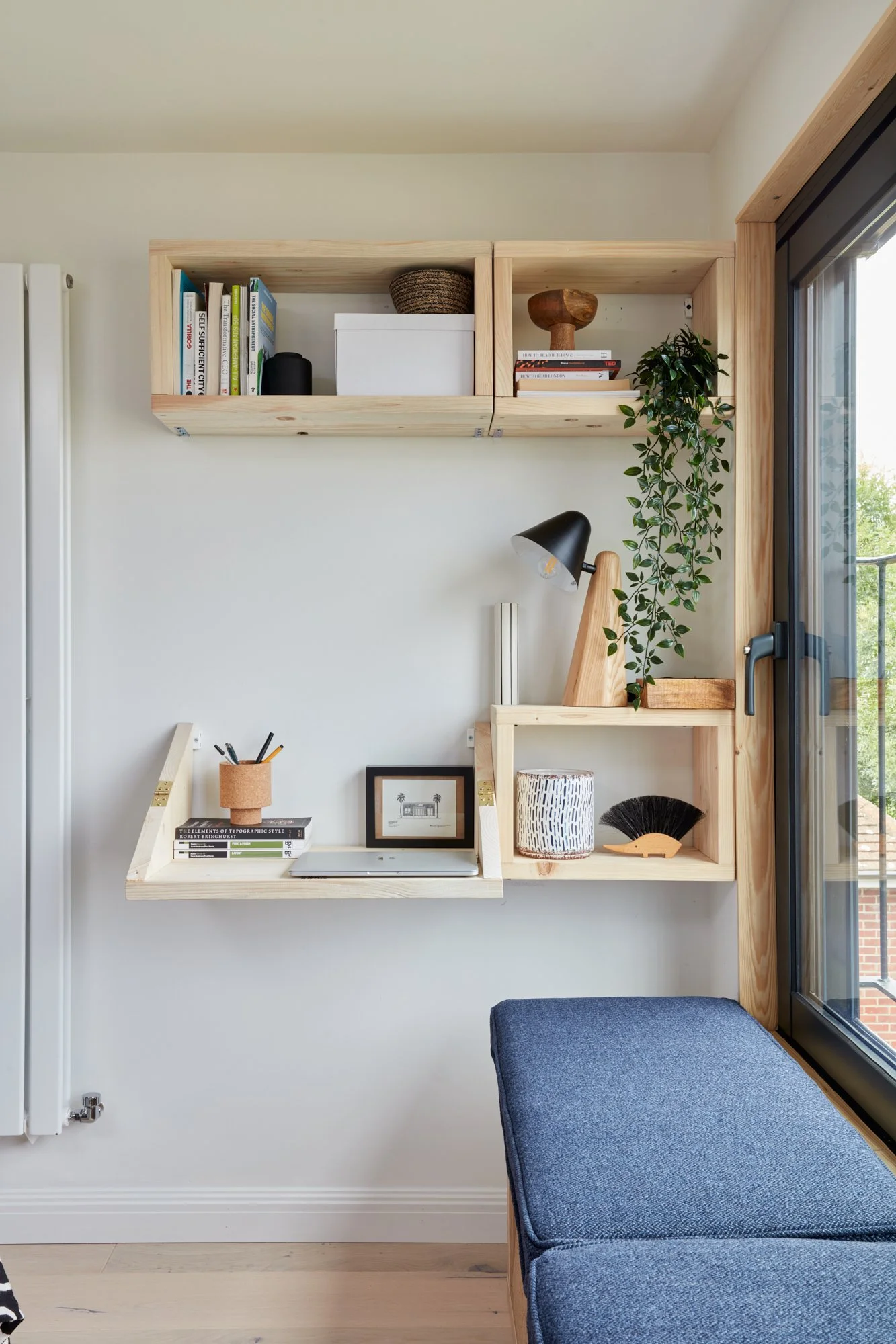 Interior of a modern room with wooden floating shelves, a small desk with a laptop, framed picture, and decorative objects, next to a large window with a blue upholstered bench in the foreground.
