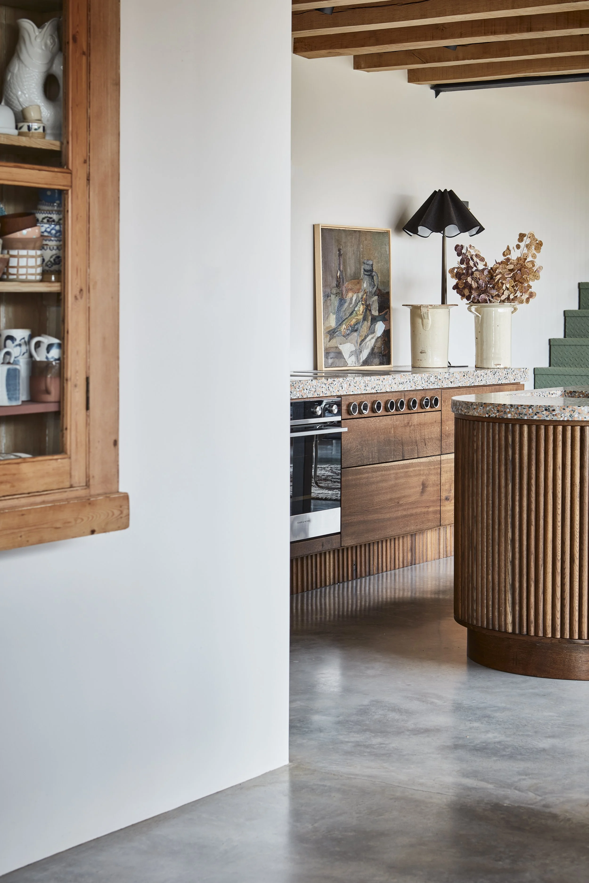 Part of a kitchen interior with a wooden cabinet on the left, a white wall, a painting, two cream-colored vases with dried flowers, a black table lamp, and green seating in the background.