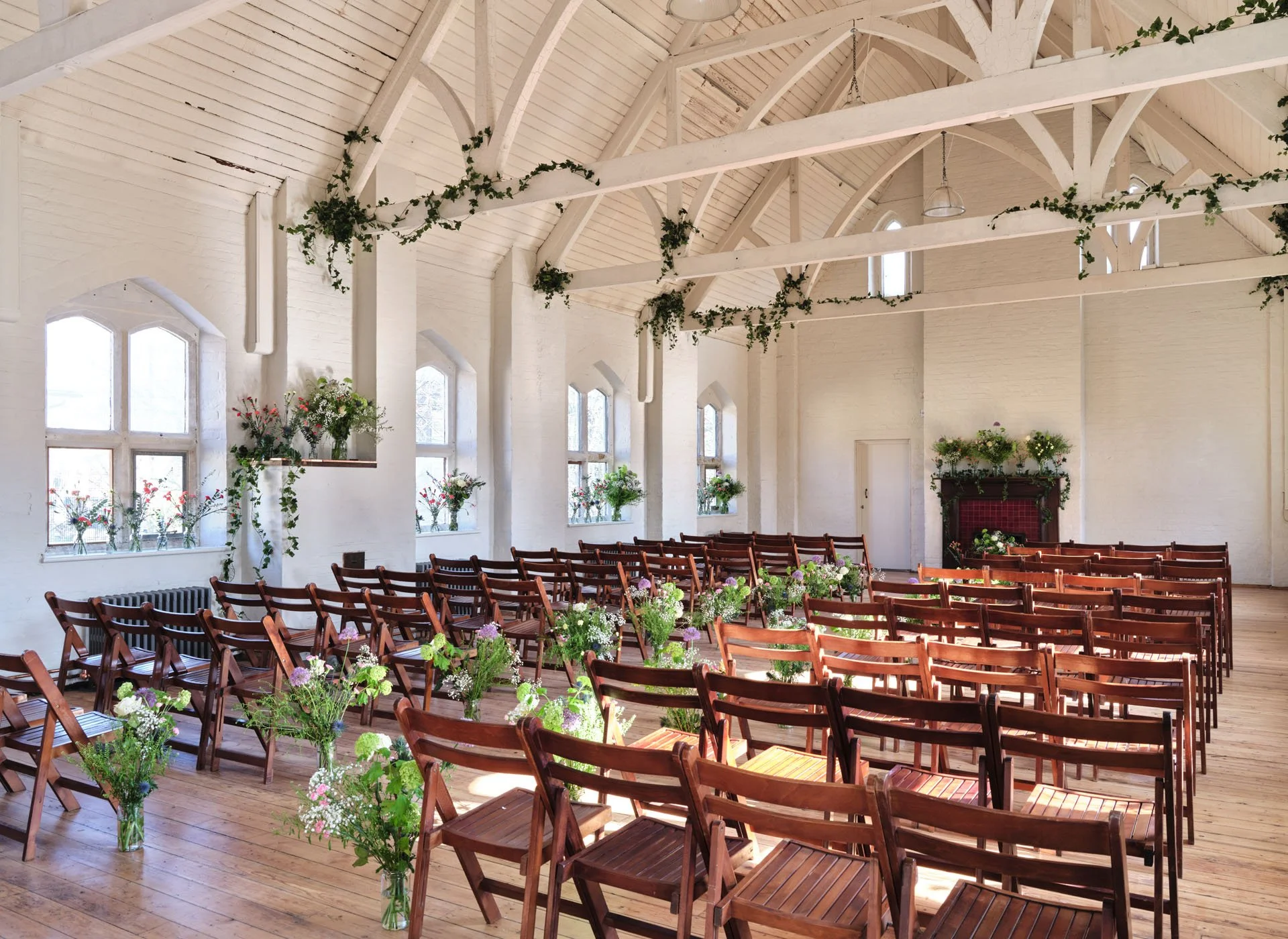 Indoor wedding ceremony setup with wooden chairs, floral arrangements in vases, and natural light streaming through large arched windows in a white-walled, high-beamed ceiling room.