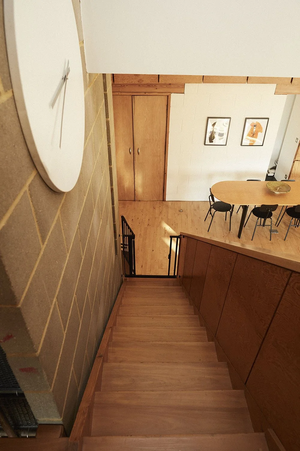 View of a wooden staircase leading down to a dining area with a wooden table and black chairs, framed artwork on the wall, and a wooden closet door, with a brick wall and a clock visible.