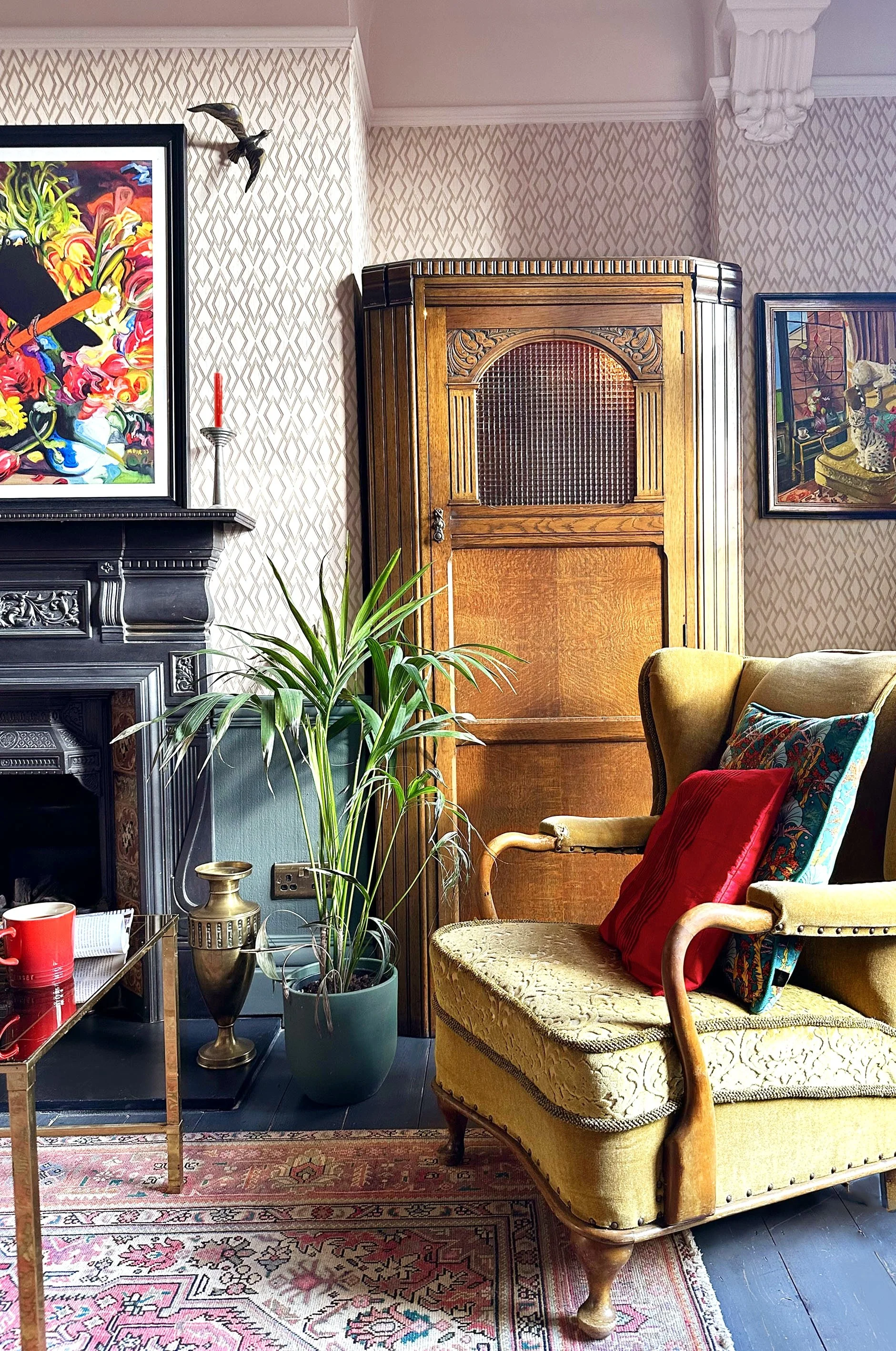 A cozy living room corner featuring a vintage yellow upholstered armchair with pillows, a green potted plant, a black fireplace with intricate detailing, a wooden cabinet, colorful artwork, a gold vase, and a small table with a red mug. The room has 