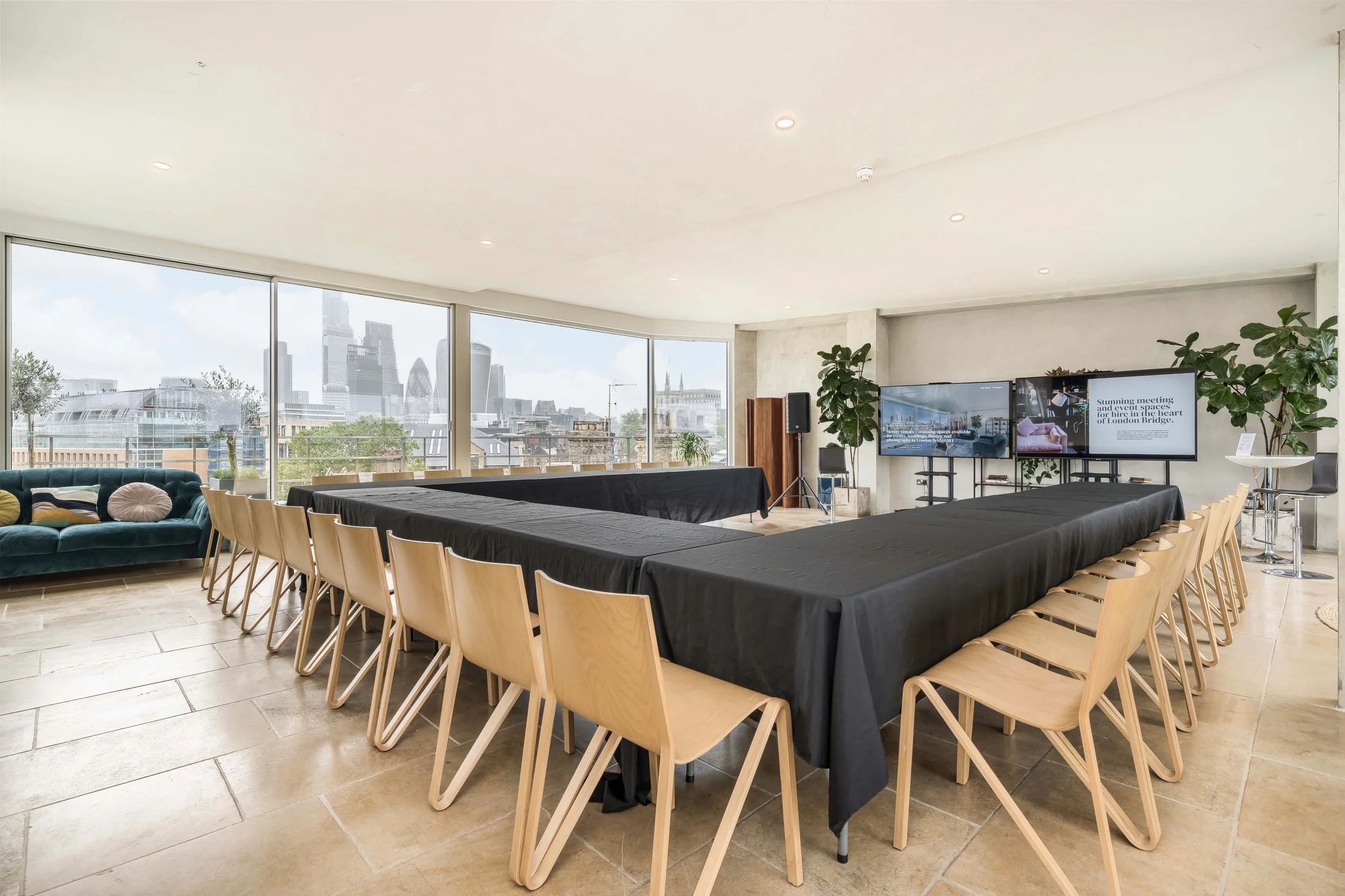 Modern conference room with large windows showing a city skyline, arranged with long tables draped in black cloth, surrounded by wooden chairs, and a television screen at the front of the room.