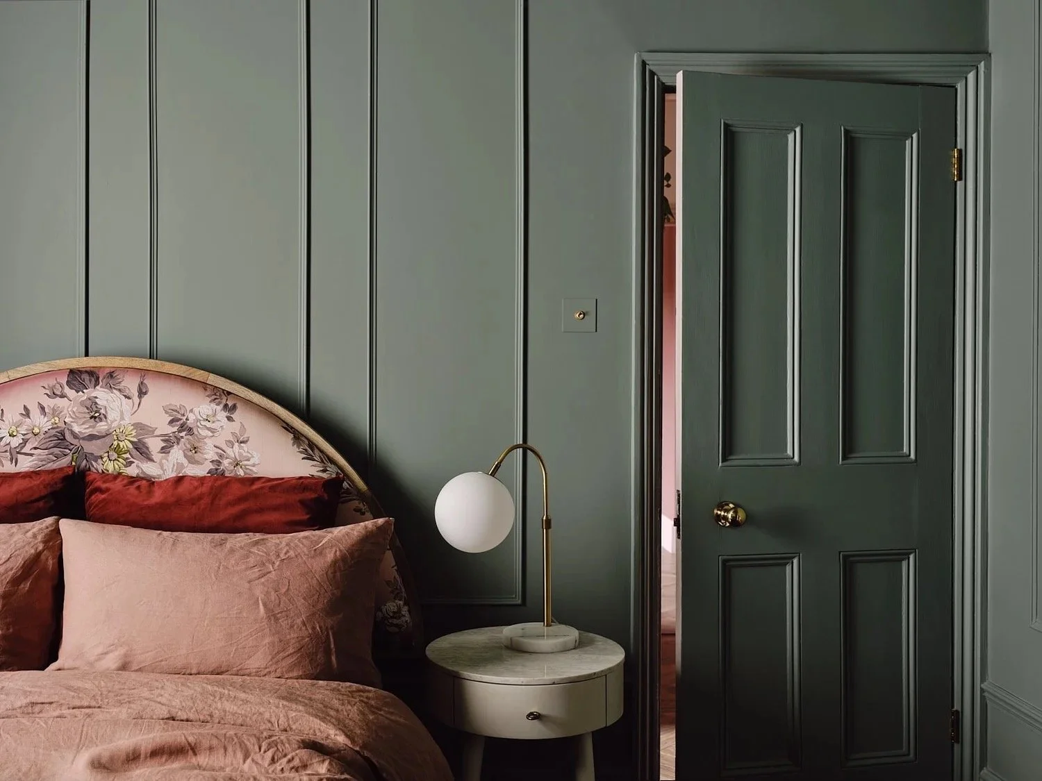 A bedroom with a dark green paneled wall, a matching closed door, a floral upholstered headboard, peach and burgundy pillows, and a white bedside table with a modern white and brass lamp