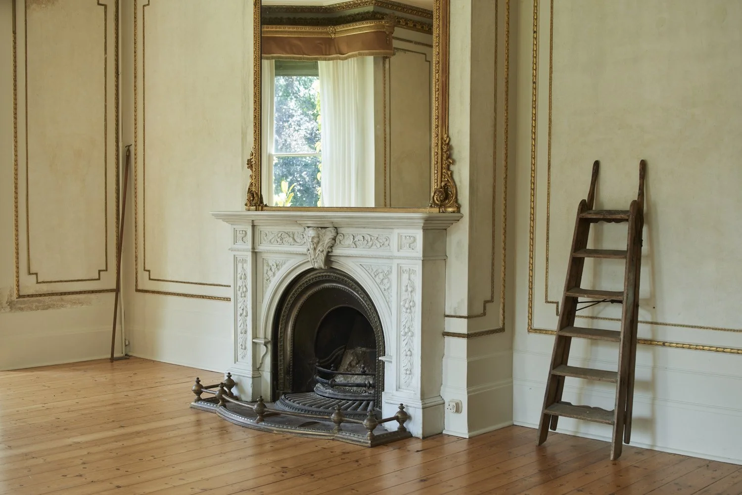 An elegant room with a ornate white fireplace, a large mirror above, and a wooden ladder leaning against the wall. The room has cream-colored walls with gold trim and a hardwood floor. A window with sheer curtains lets in natural light, revealing gre