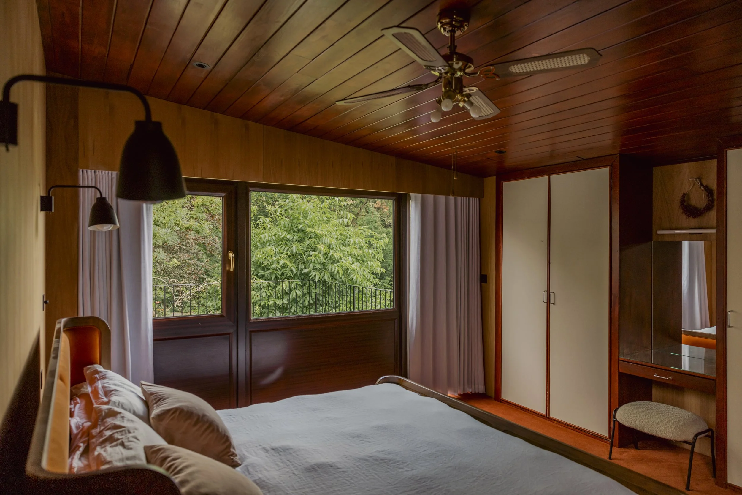 A cozy bedroom with wood-paneled ceiling and walls, large window showing green trees outside, a bed with white bedding and pillows, a ceiling fan, and a built-in wardrobe with sliding doors.