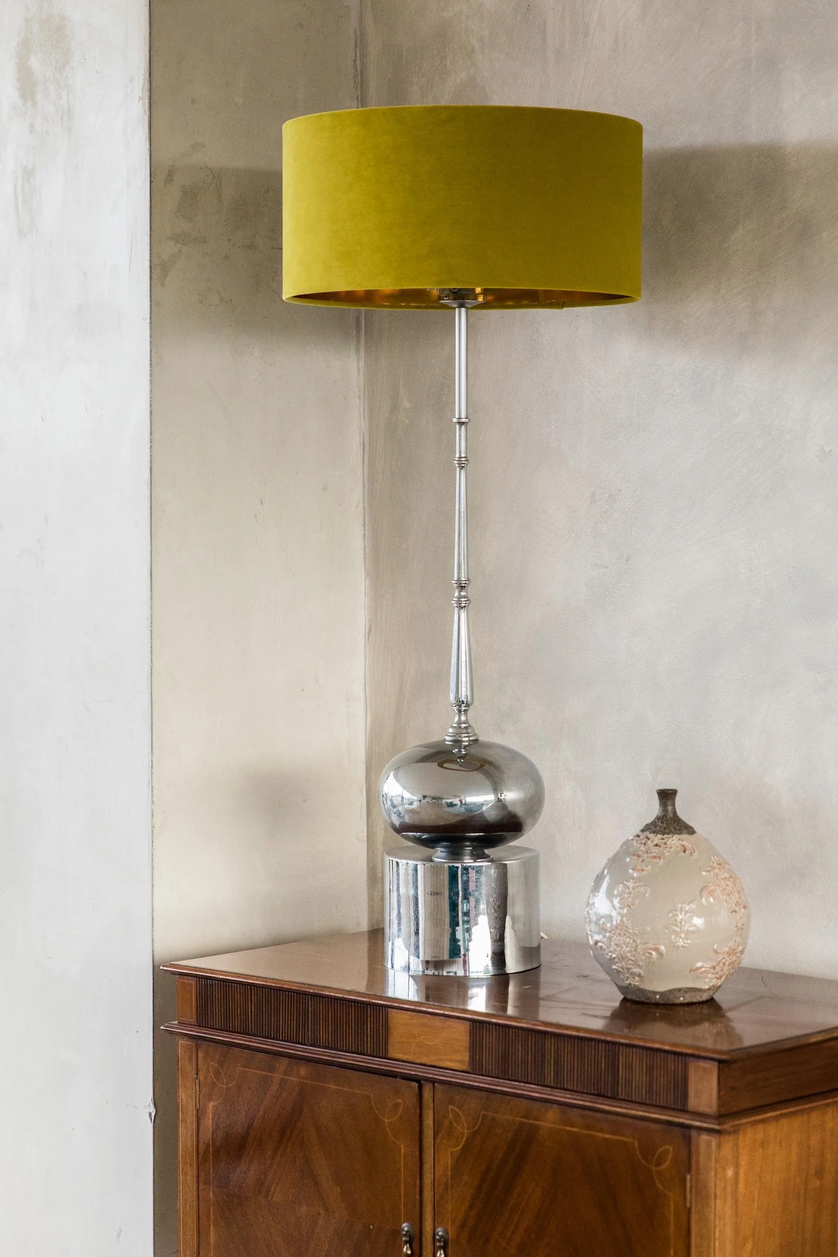 A decorative table lamp with a shiny silver base and a tall, slender stem, topped with an olive green lampshade, is placed on a wooden cabinet next to a frosted glass vase with floral etchings.