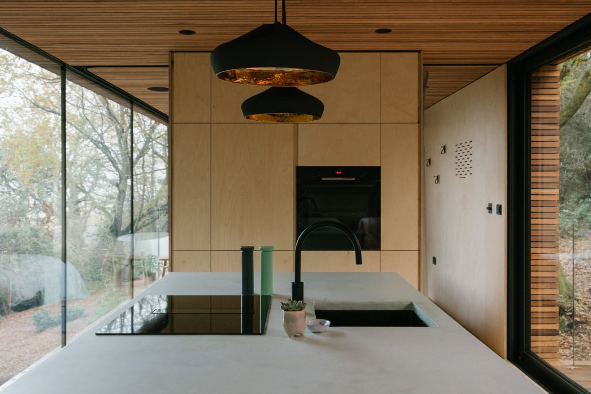 Modern kitchen with a white countertop, black sink, black faucet, wood cabinets, and large glass windows showing outdoor trees.