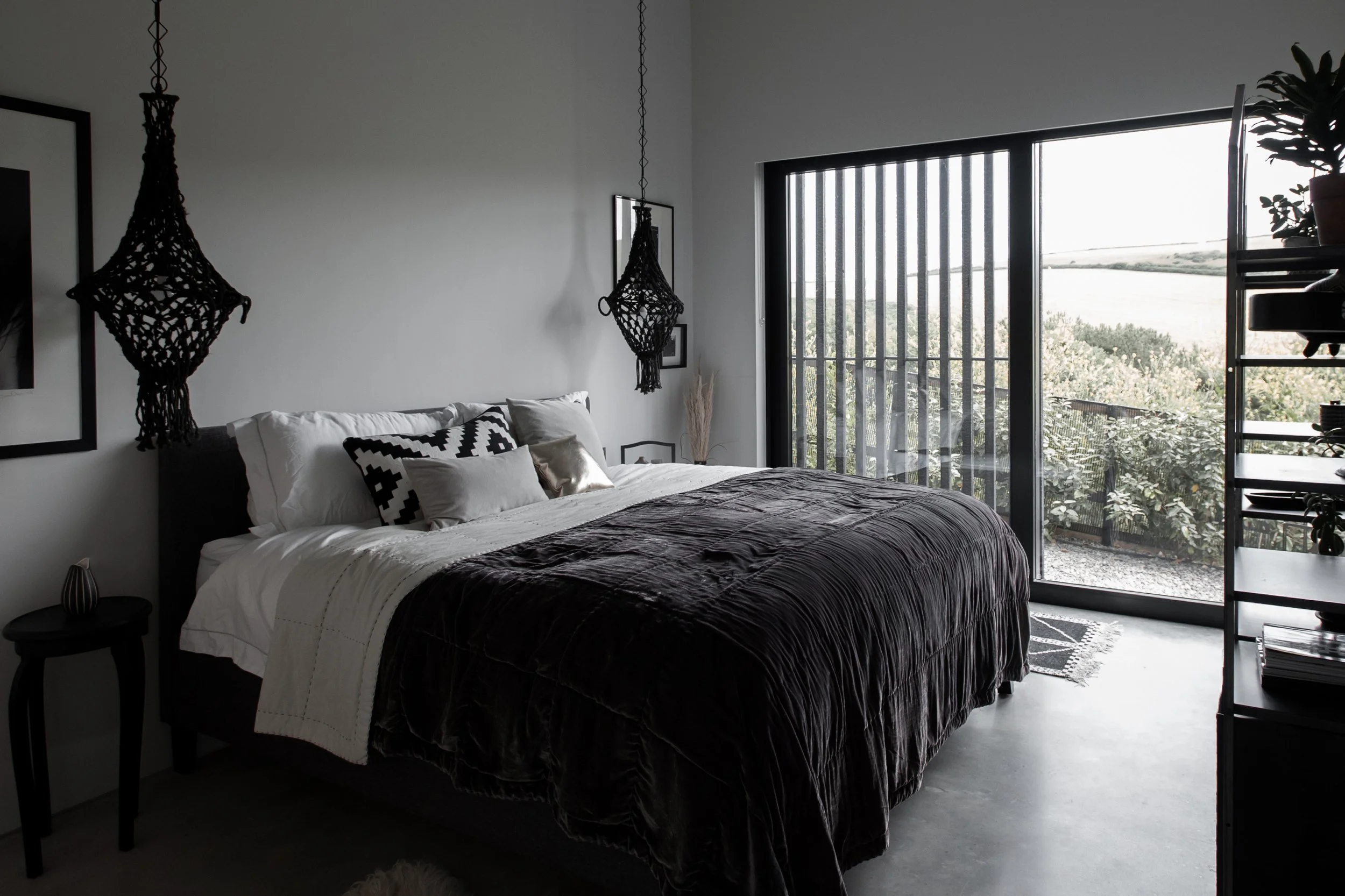 Modern bedroom with black and white decor, large window with black bars overlooking greenery, black side table, and hanging black macramé decorations.