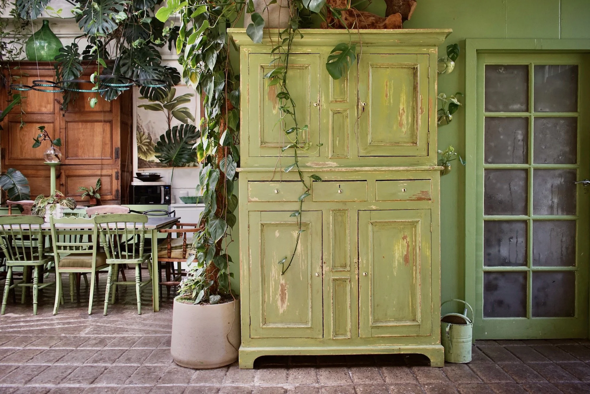 Green distressed cabinet next to a glass-paned door in a rustic interior with wooden furniture, green chairs, potted plants, and a brick floor.