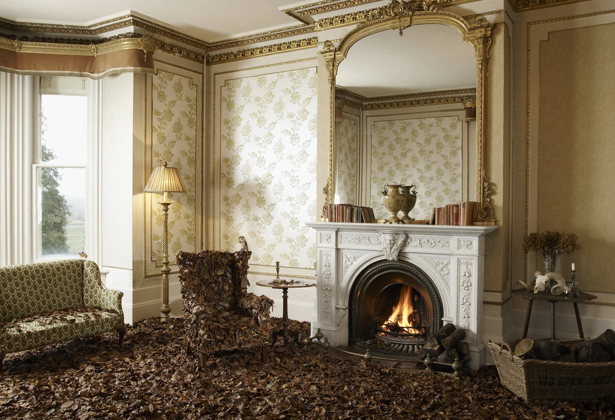 Living room with ornate gold trim, floral wallpaper, and a white fireplace with a fire burning. The floor is covered in fallen leaves. There is an antique-style green patterned sofa, a tall floor lamp, a chair covered in dried leaves, and a side tabl