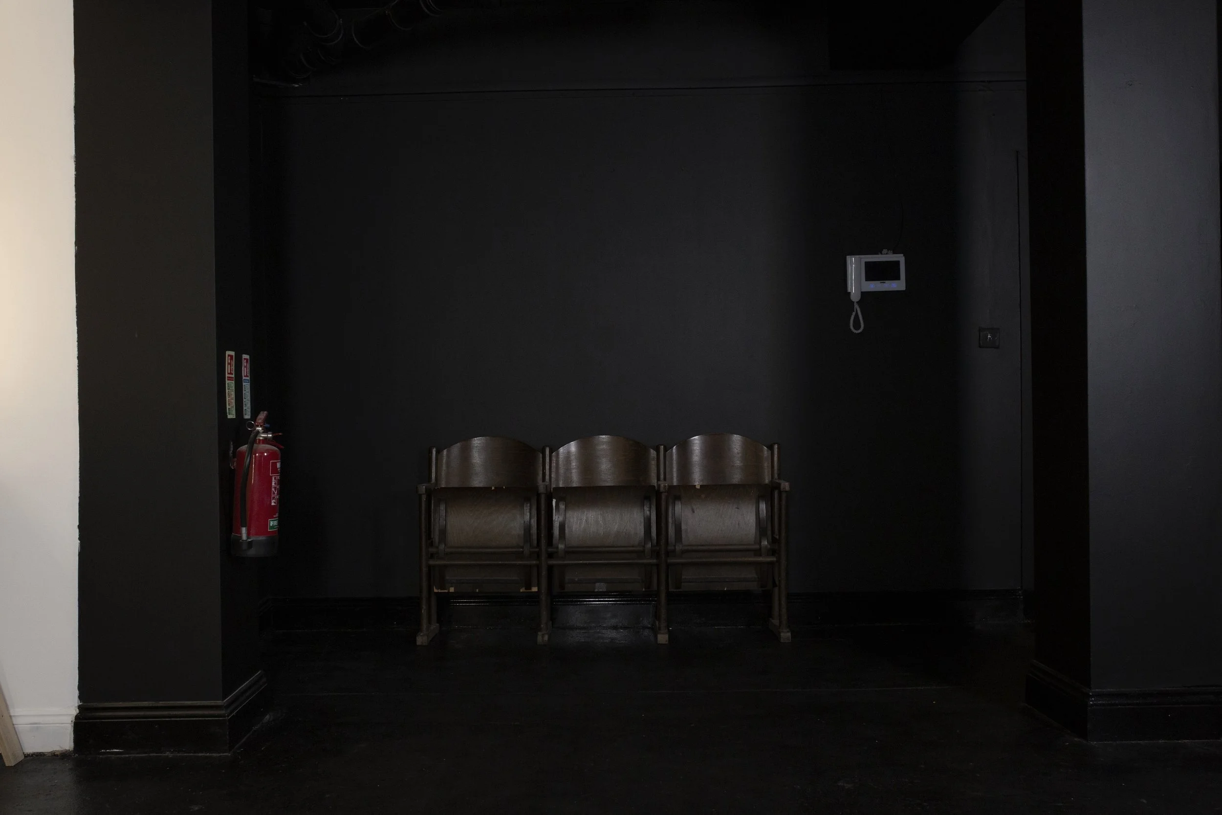 Three wooden chairs lined up against a black wall with a fire extinguisher on the left and a wall-mounted intercom or phone on the right.