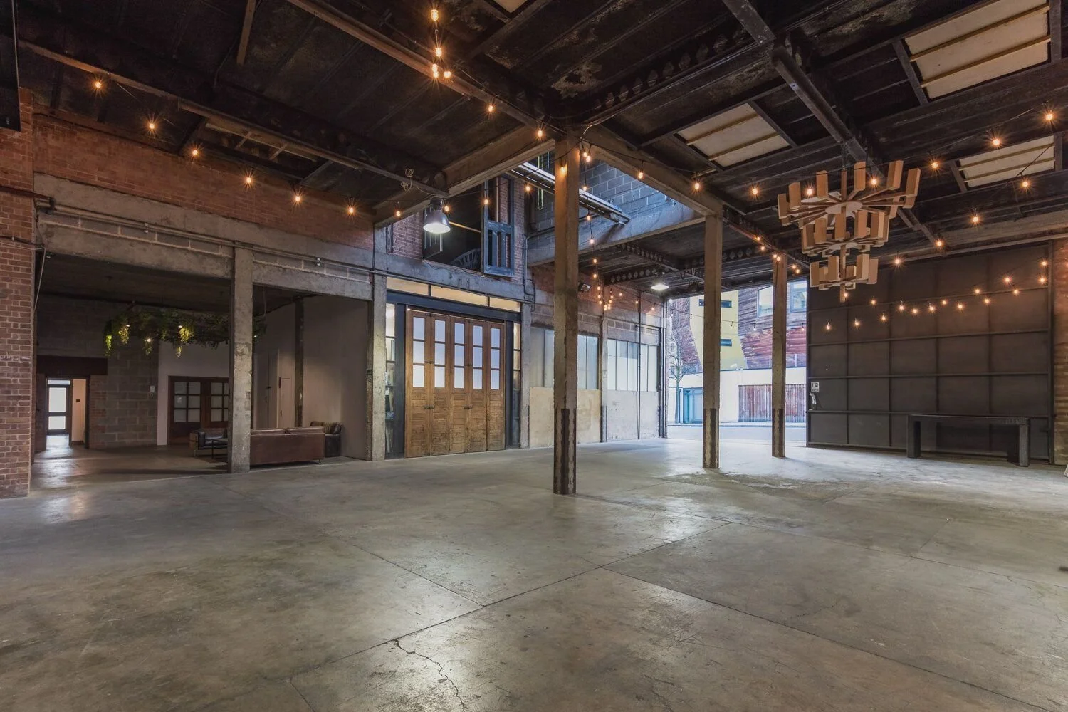 Interior of an industrial-style building with exposed brick and concrete walls, string lights hanging from the ceiling, wooden and metal accents, a chandelier, and open space with some furniture in the background.