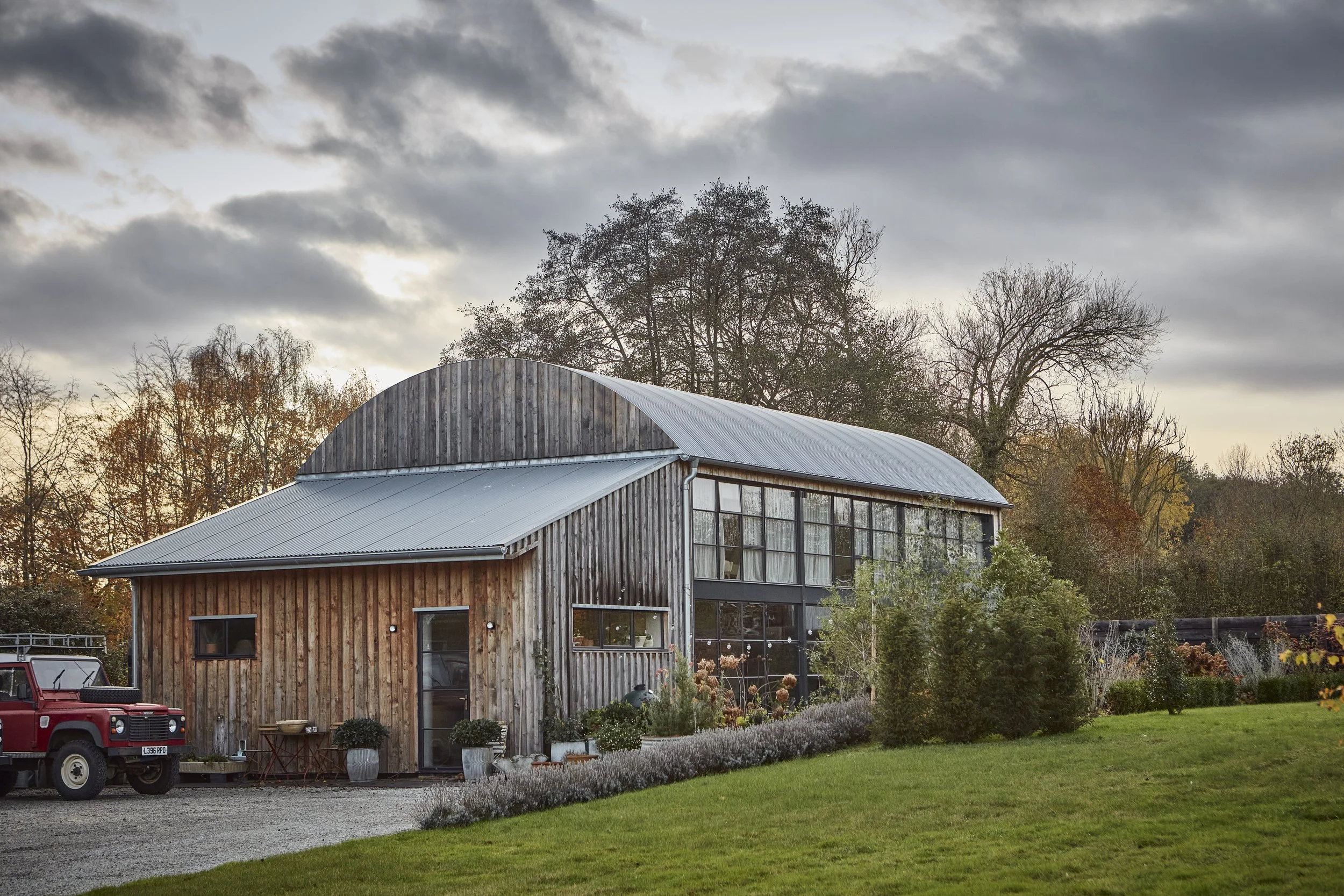 A large wooden and metal barn or shed with a curved roof, surrounded by trees with fall foliage and a well-maintained lawn. A red pickup truck is parked nearby, with gravel and garden pots in the foreground.