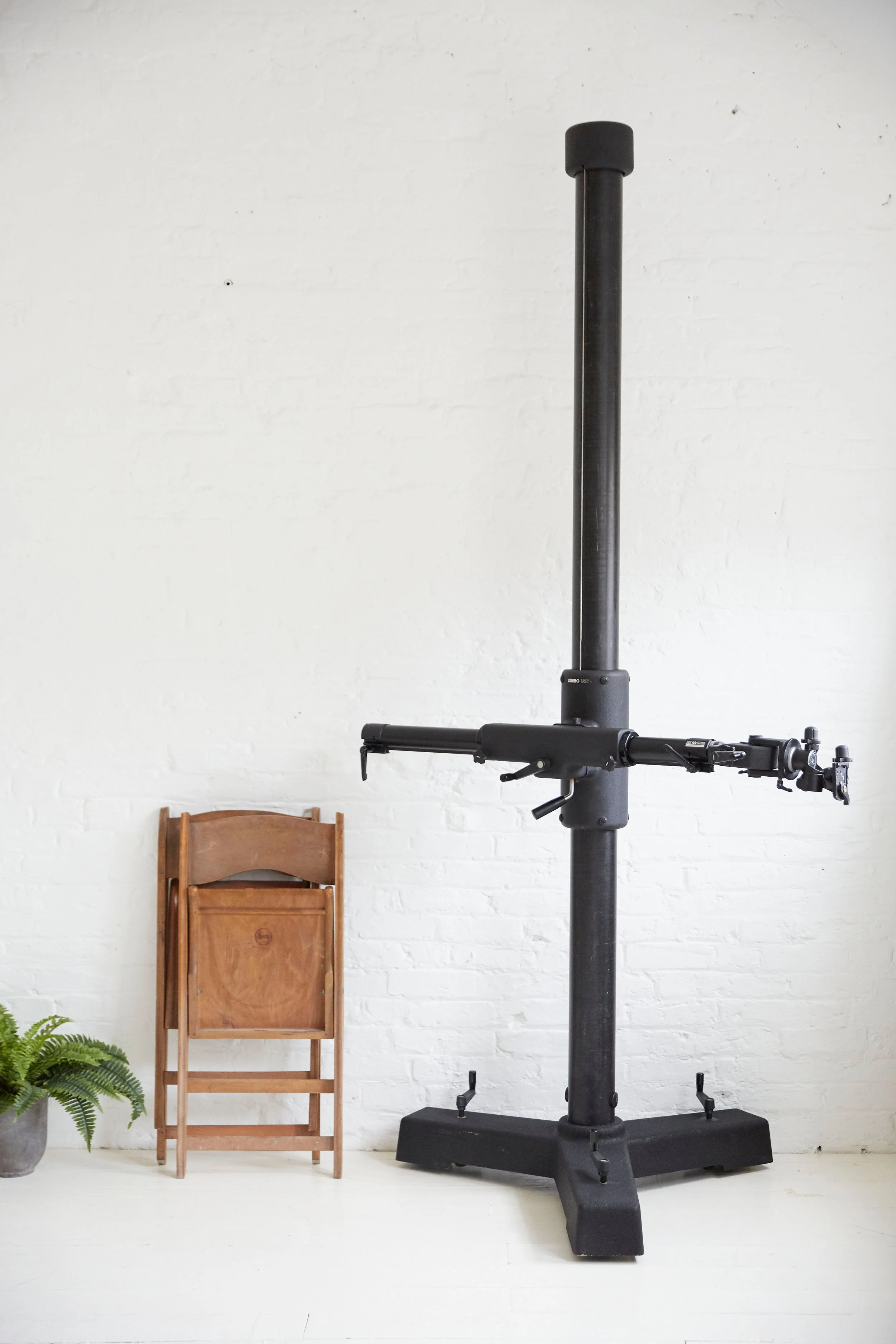 A black indoor cycling trainer stand on a white floor with a white wall in the background, next to a small potted green fern and a folded wooden chair.