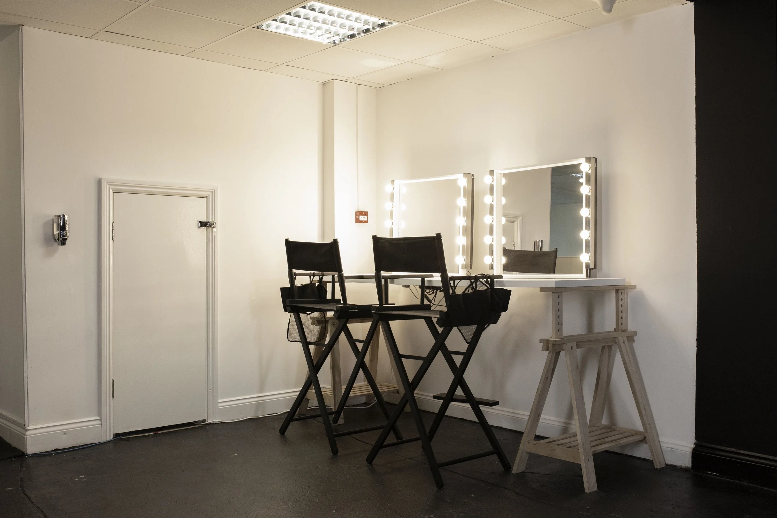 A vanity table with two mirrors and unlit lights, two high black chairs, and a small white table on a black and white floor in a room with a white and black wall.