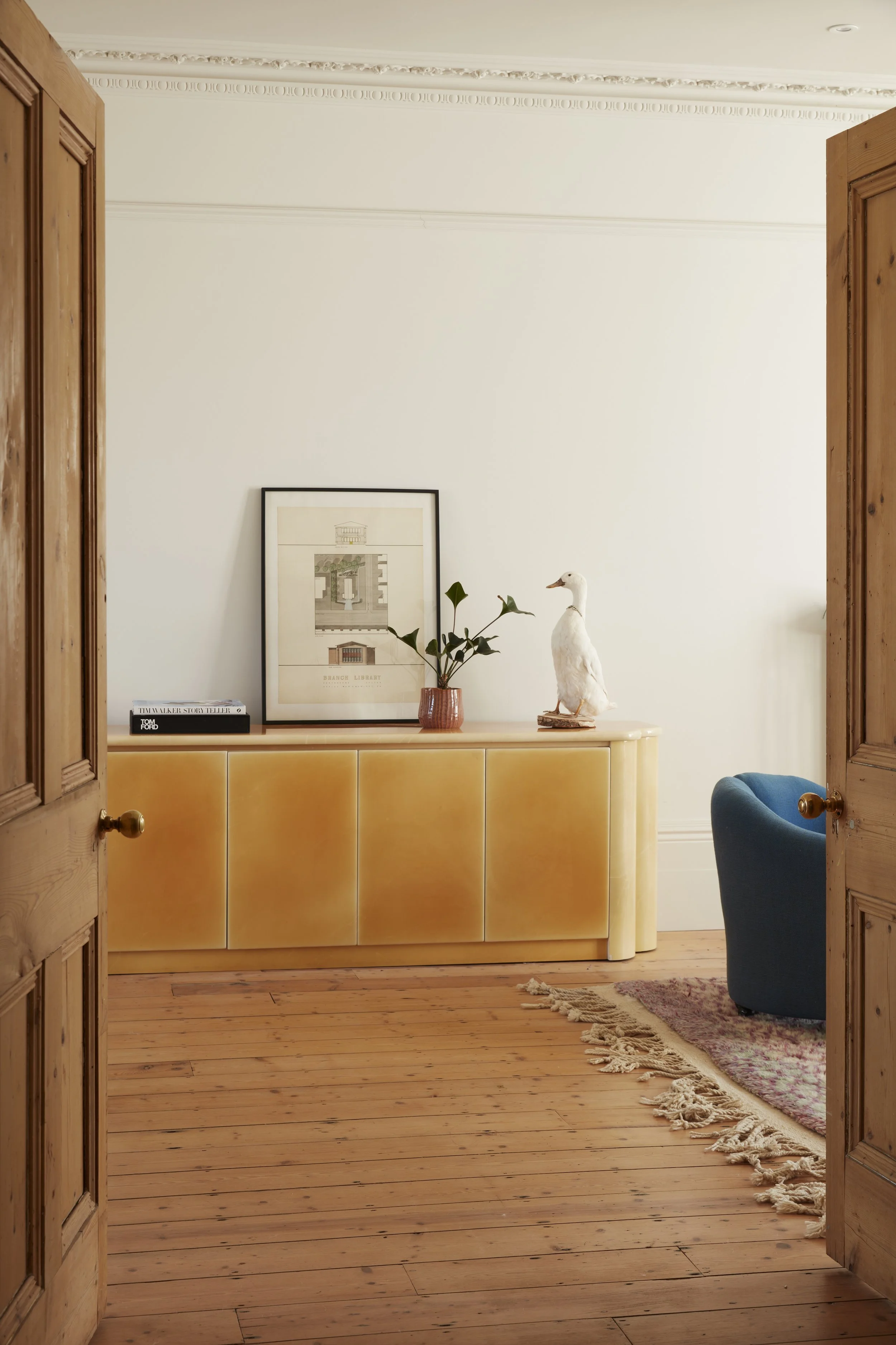 View through wooden doors into a room with a yellow sideboard, a framed artwork, a potted plant, and a white duck figurine on top. There is a blue armchair and a fringe rug on a wooden floor.