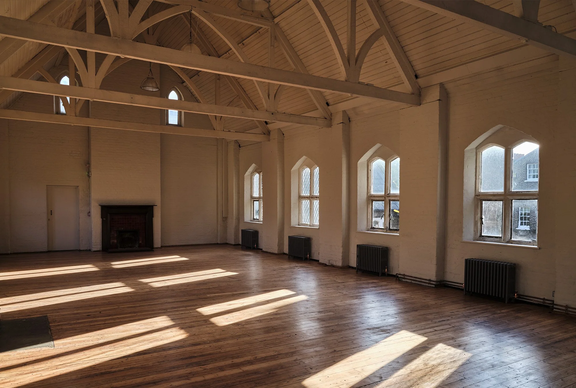 Empty large room with wooden floors, white brick walls, large arched windows, and exposed ceiling beams, with sunlight streaming in.