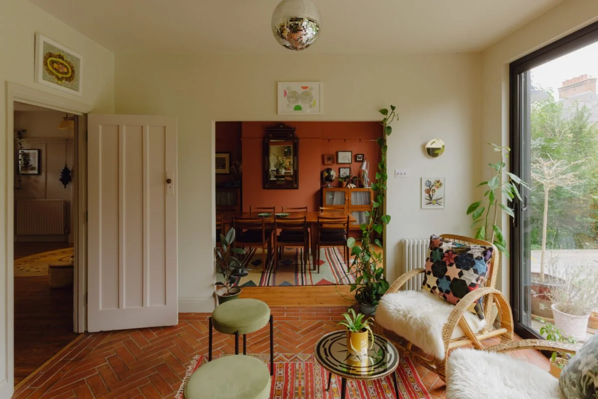 A bright living room with large window, plants, chairs, and a glass table, connecting to a dining area with a table and chairs and artwork on the walls.