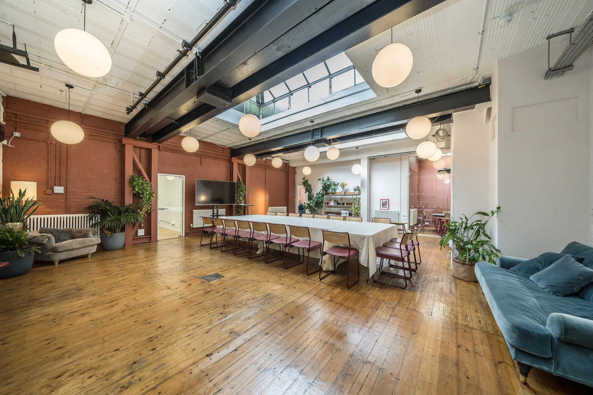 Bright, spacious room with wooden floors, white ceiling, large skylight, round pendant lights, green potted plants, a long conference table with chairs, a blue velvet sofa, a large TV, and a bookshelf in the background.