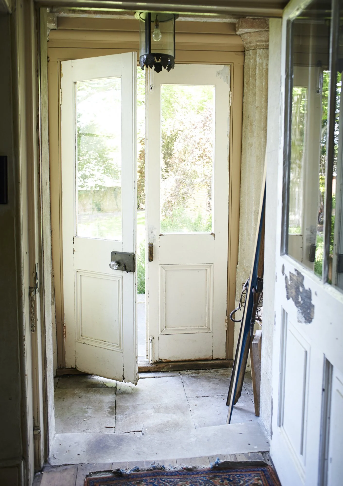 An entryway with partially open, weathered white double doors leading outside to a green garden. The floor is tiled with some visible cracks, and there is a small area rug at the bottom of the door. The ceiling has an old light fixture hanging with n