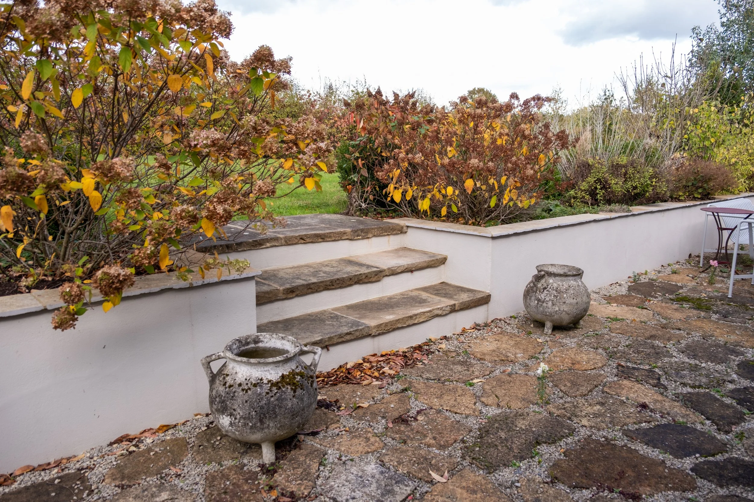 A backyard patio area with stone steps leading up to a garden with bushes and trees, some with fall foliage. There are two weathered, large, round stone planters in the foreground and a patio table with chairs to the right.