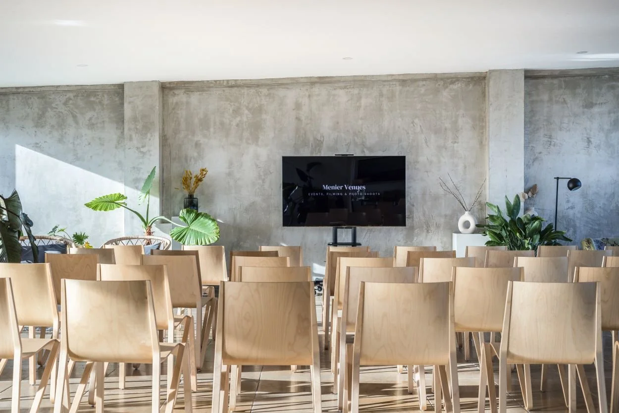 Empty conference or event room with wooden chairs facing a large flat-screen TV, concrete wall background, and potted green plants and decorative vases.