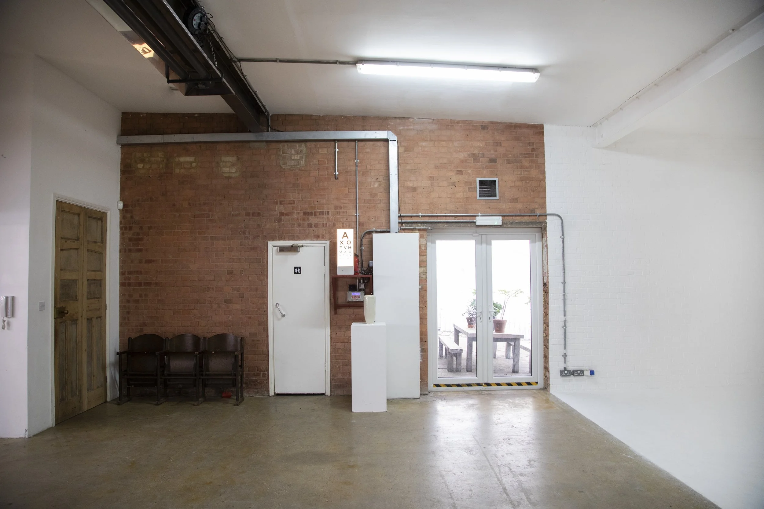 Interior of an industrial-style room with exposed brick wall, white painted wall, fluorescent ceiling lights, wooden door, and glass doors leading to an outdoor patio with plants.