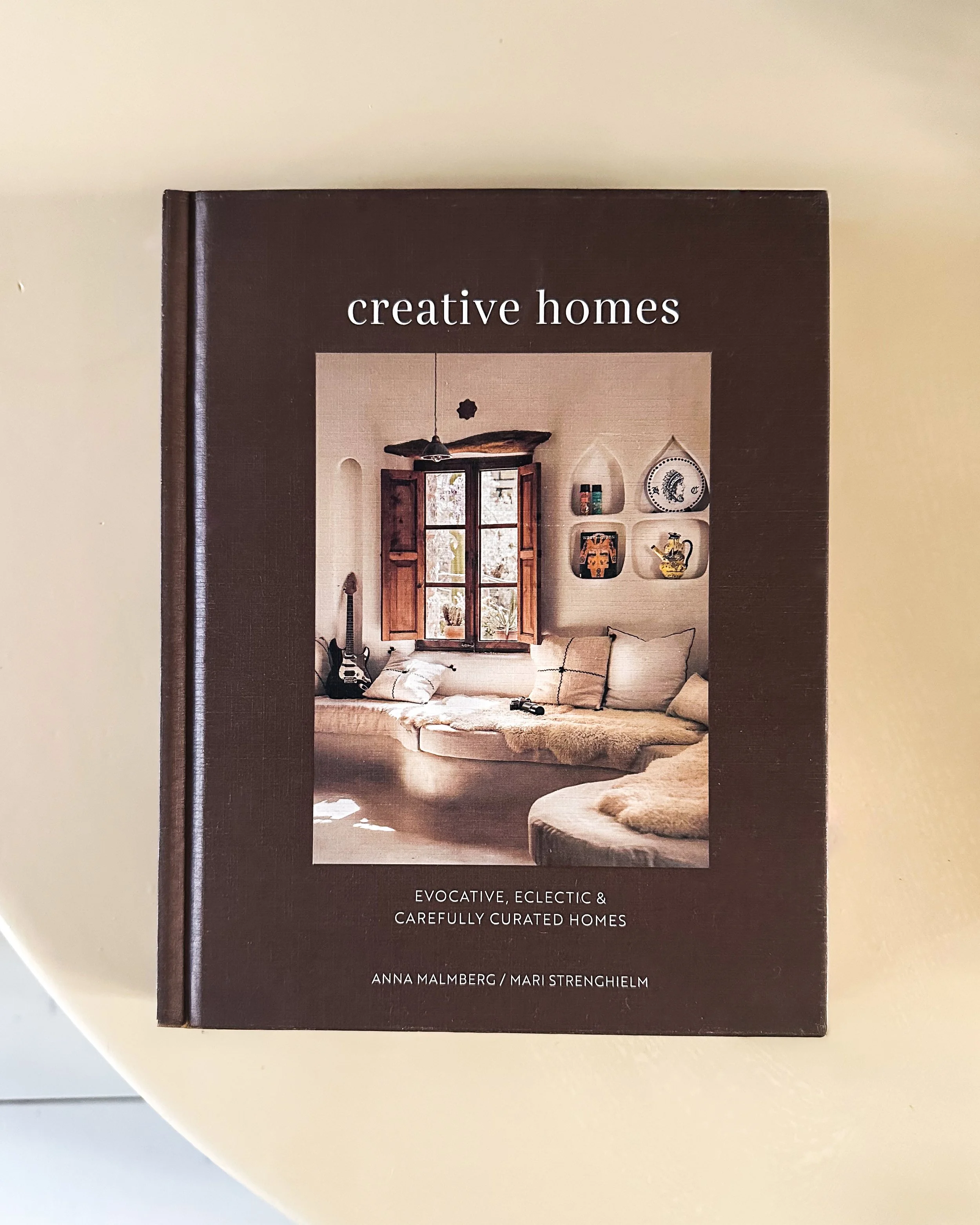 Book titled 'Creative Homes' featuring a cozy interior with a sofa, window, guitar, and shelves with decorative items.